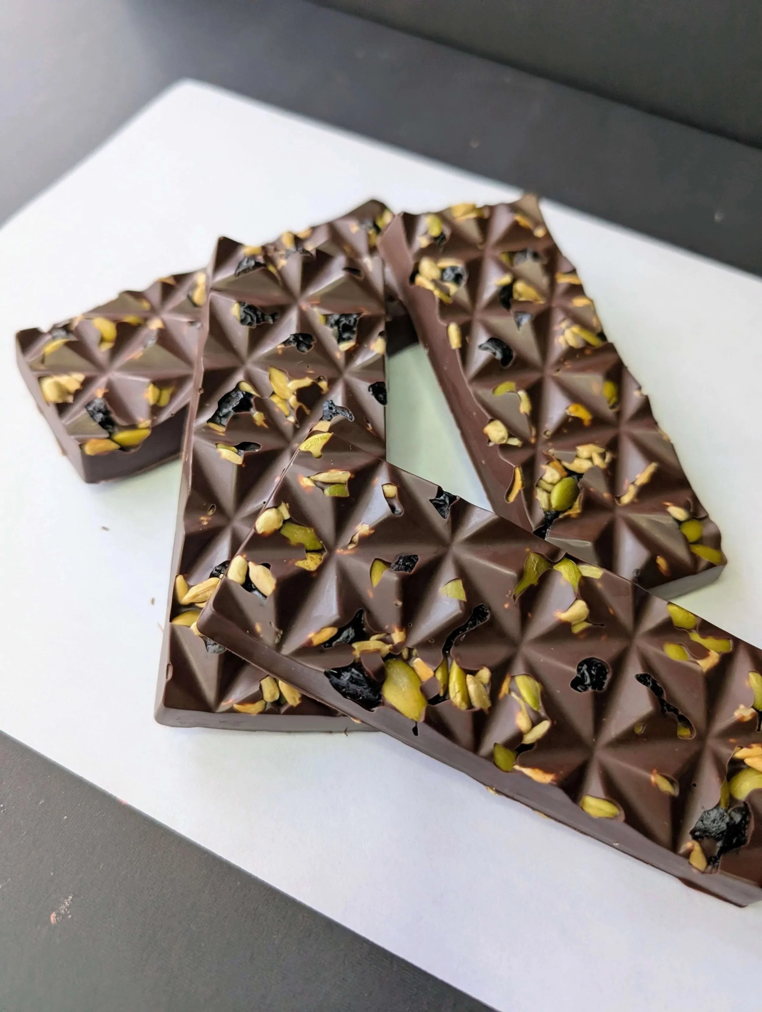 A close-up of a piece of dark chocolate with nuts and dried fruit on top, placed on a white surface.
