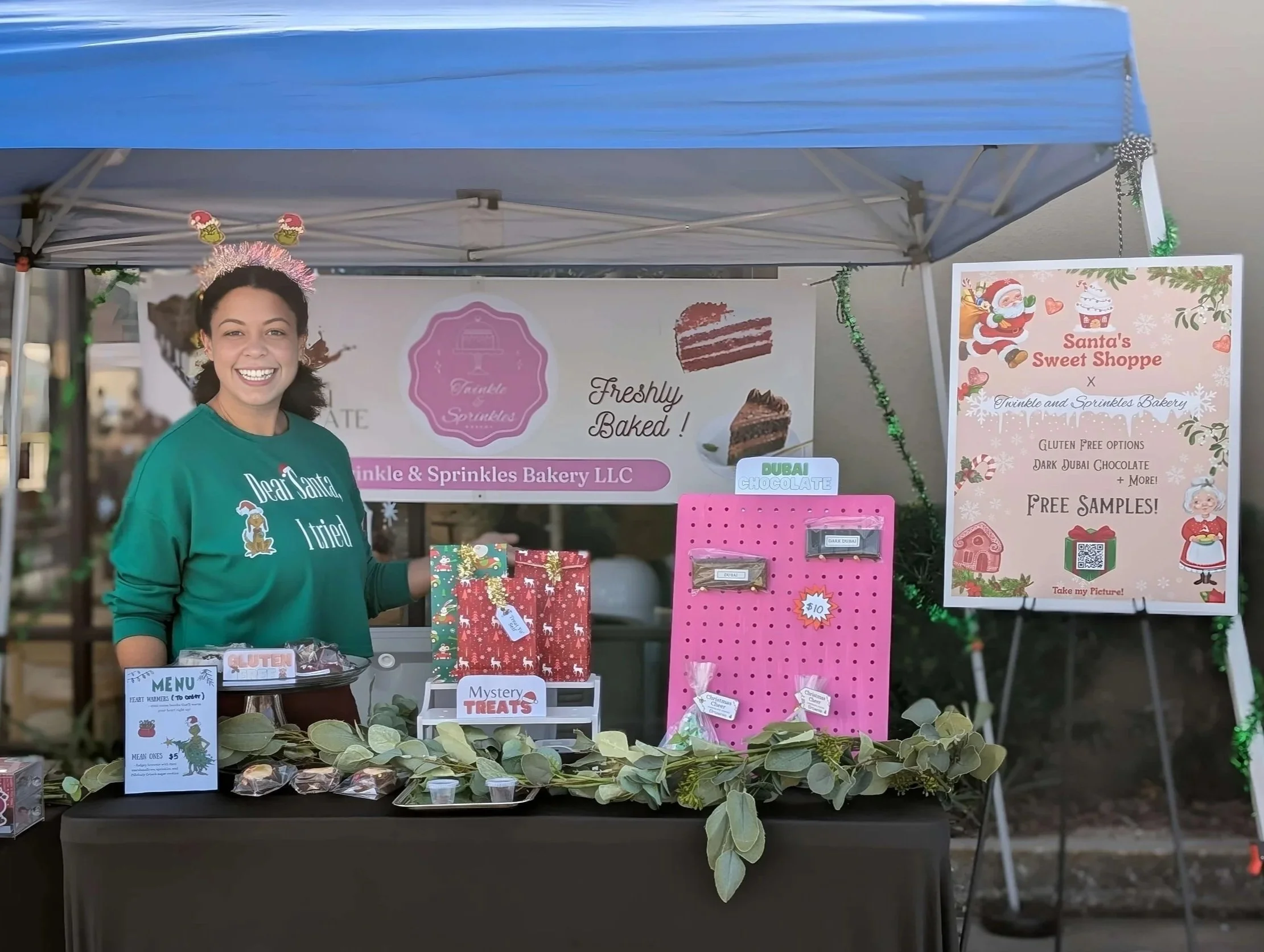 A woman smiling at a Christmas-themed bakery booth called 'Twinkle & Sprinkles Bakery LLC' with a sign advertising free samples, gluten-free options, and dark Dubai chocolate, decorated with holiday decorations, and a large sign about Santa's Sweet Shoppe.