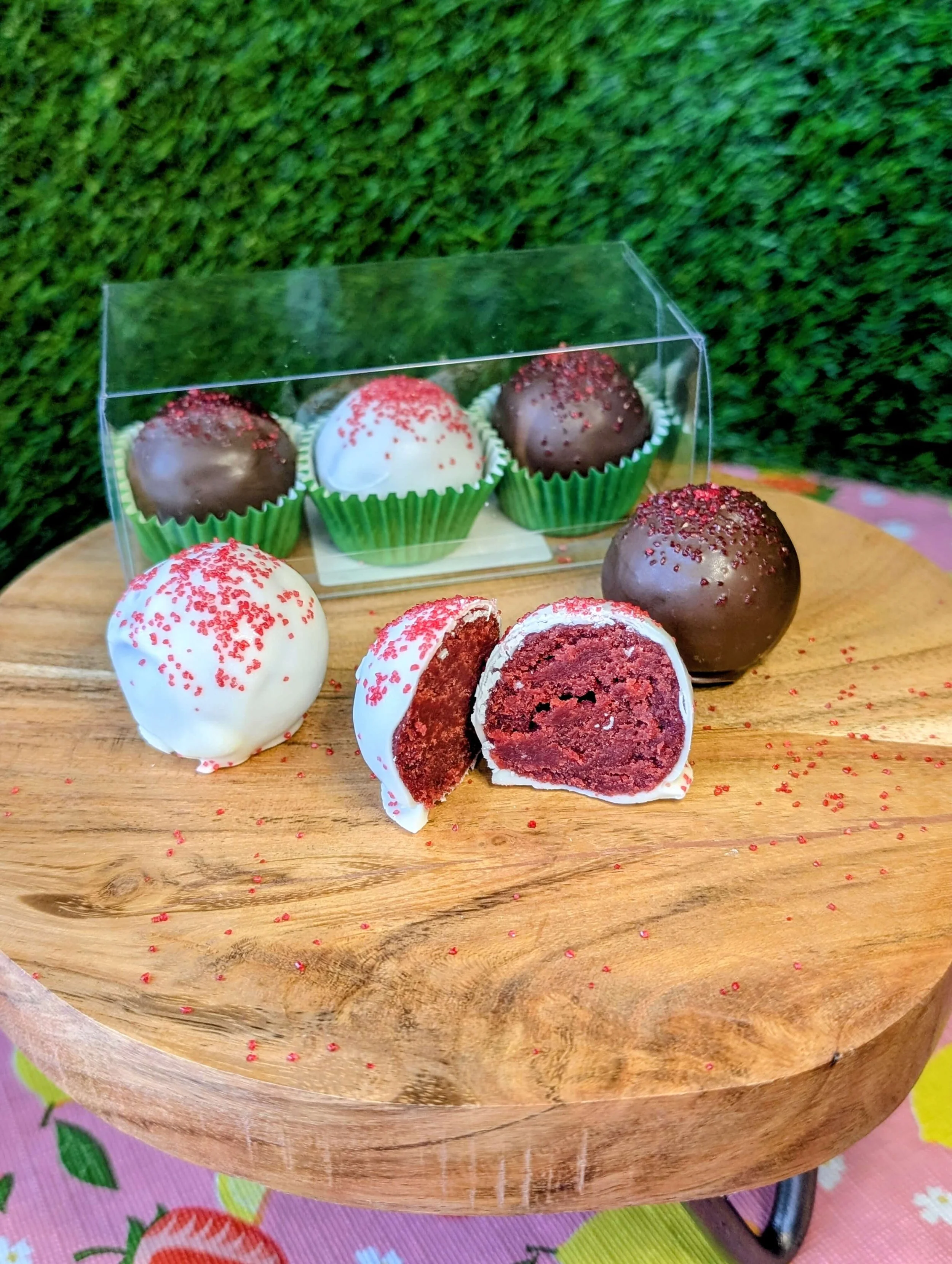 A wooden stand displays red velvet cake truffles coated in white and dark chocolate, decorated with red sprinkles. In the foreground, one white-coated truffle is cut in half, revealing a rich red velvet filling. Behind them, a clear plastic box holds