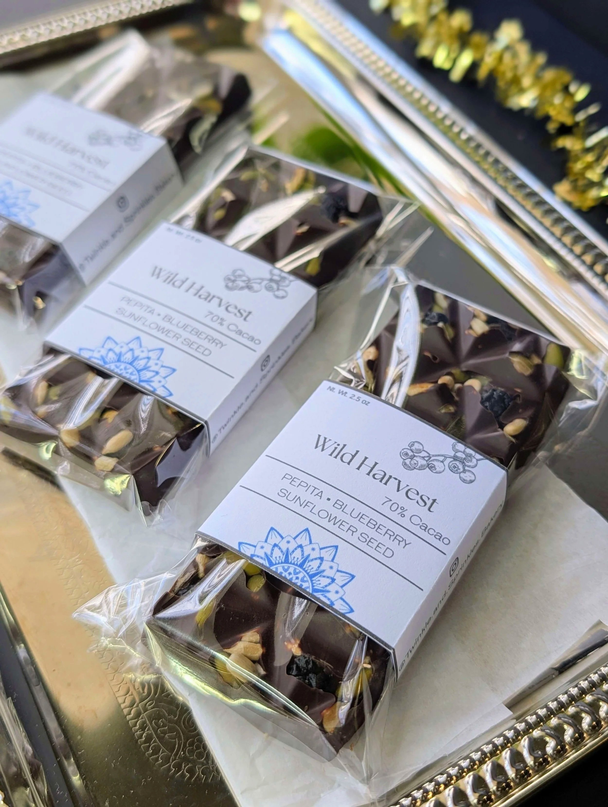 Three wrapped "Wild Harvest" 70% cacao dark chocolate bars, topped with pepitas, blueberries, and sunflower seeds, are arranged on a silver tray with gold tinsel. The white labels feature a blue mandala design.