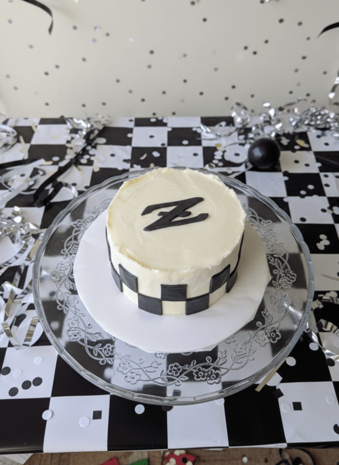 White birthday cake with black checkerboard pattern and black decorative letter on top, placed on a glass platter with a black and white checkered tablecloth and silver confetti.