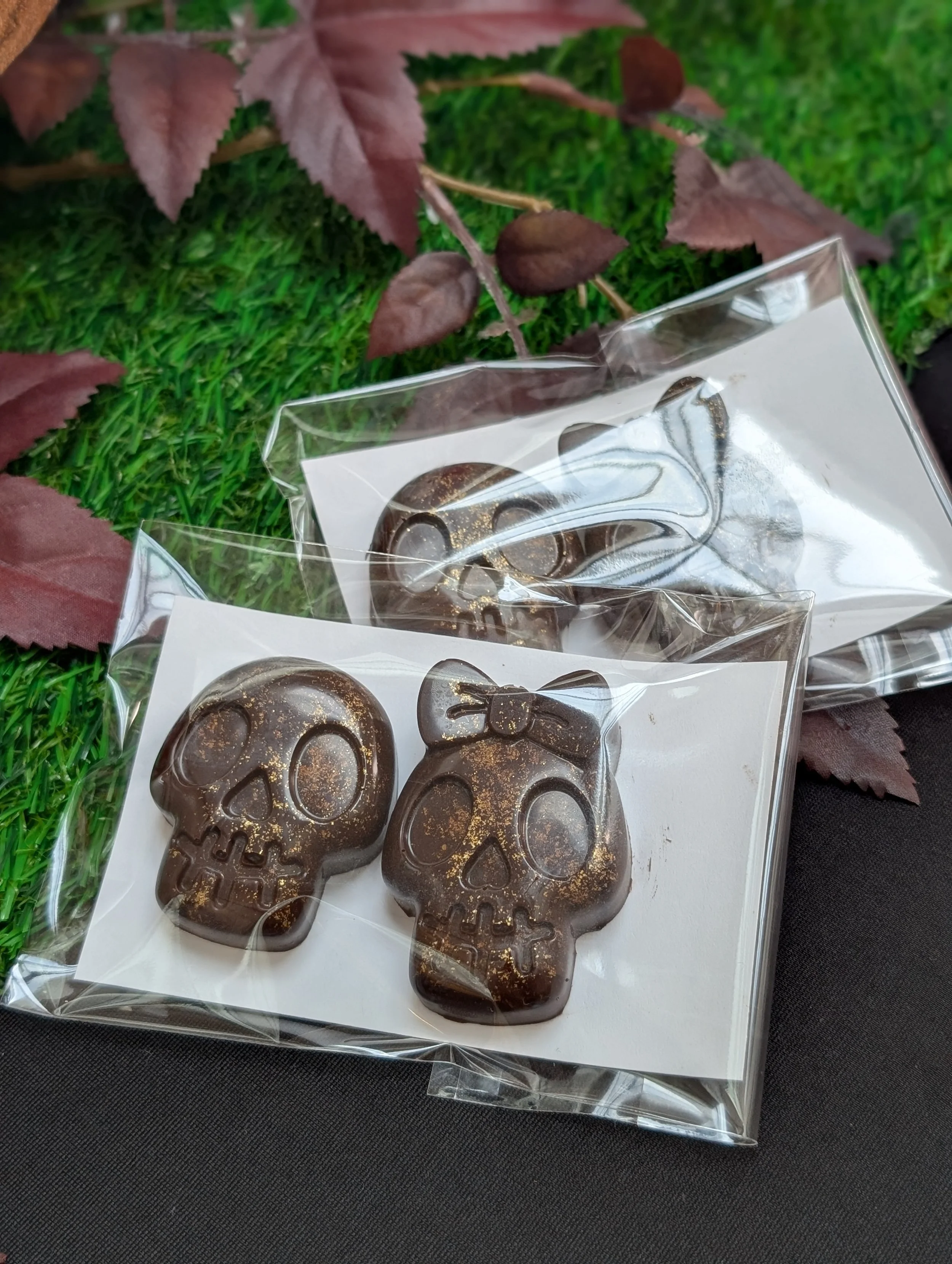 Two individually wrapped sets of dark chocolate skulls dusted with edible gold shimmer, resting on artificial grass and dark red leaves.