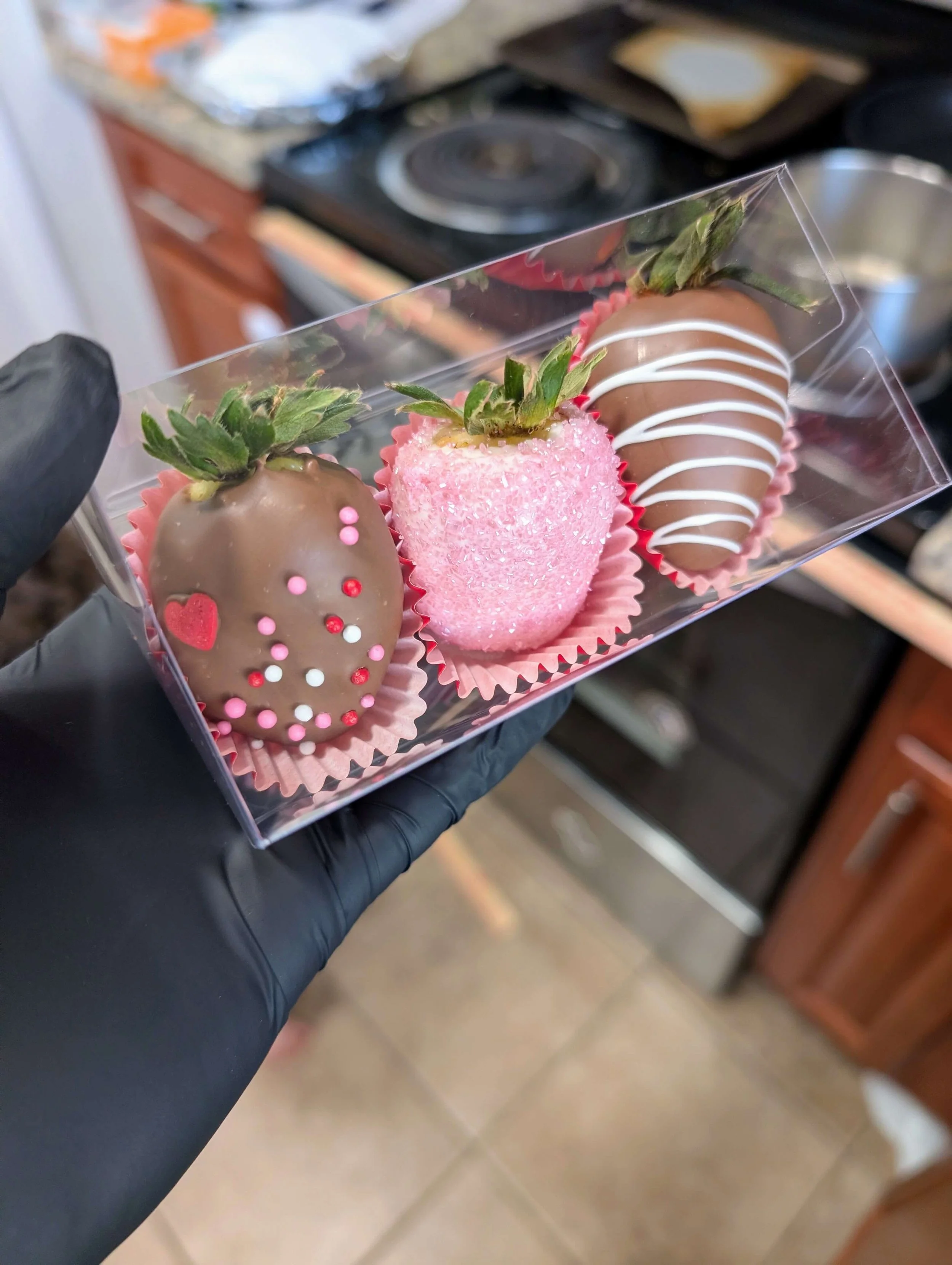 A hand wearing a black gloved hand holds a clear plastic rectangular box containing three decorated chocolate-covered strawberries in pink paper liners. The strawberry on the left is dipped in milk chocolate and decorated with pink, red, and white sp