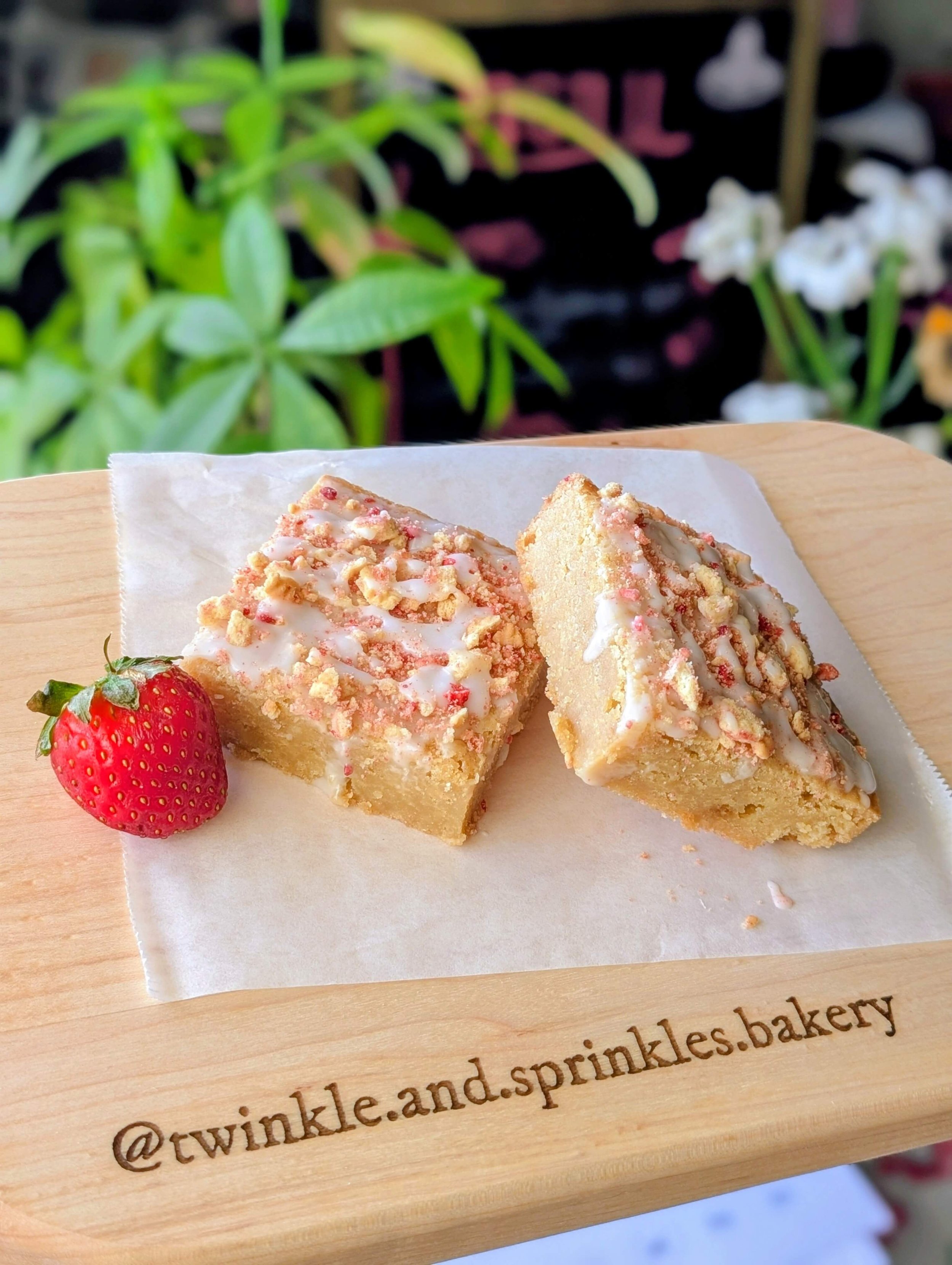 Two square strawberry shortcake blondies drizzled with white icing and topped with crumbled strawberry crunch, served next to a fresh strawberry on a wooden board engraved with "@twinkle.and.sprinkles.bakery", with a blurred money tree plant in the b