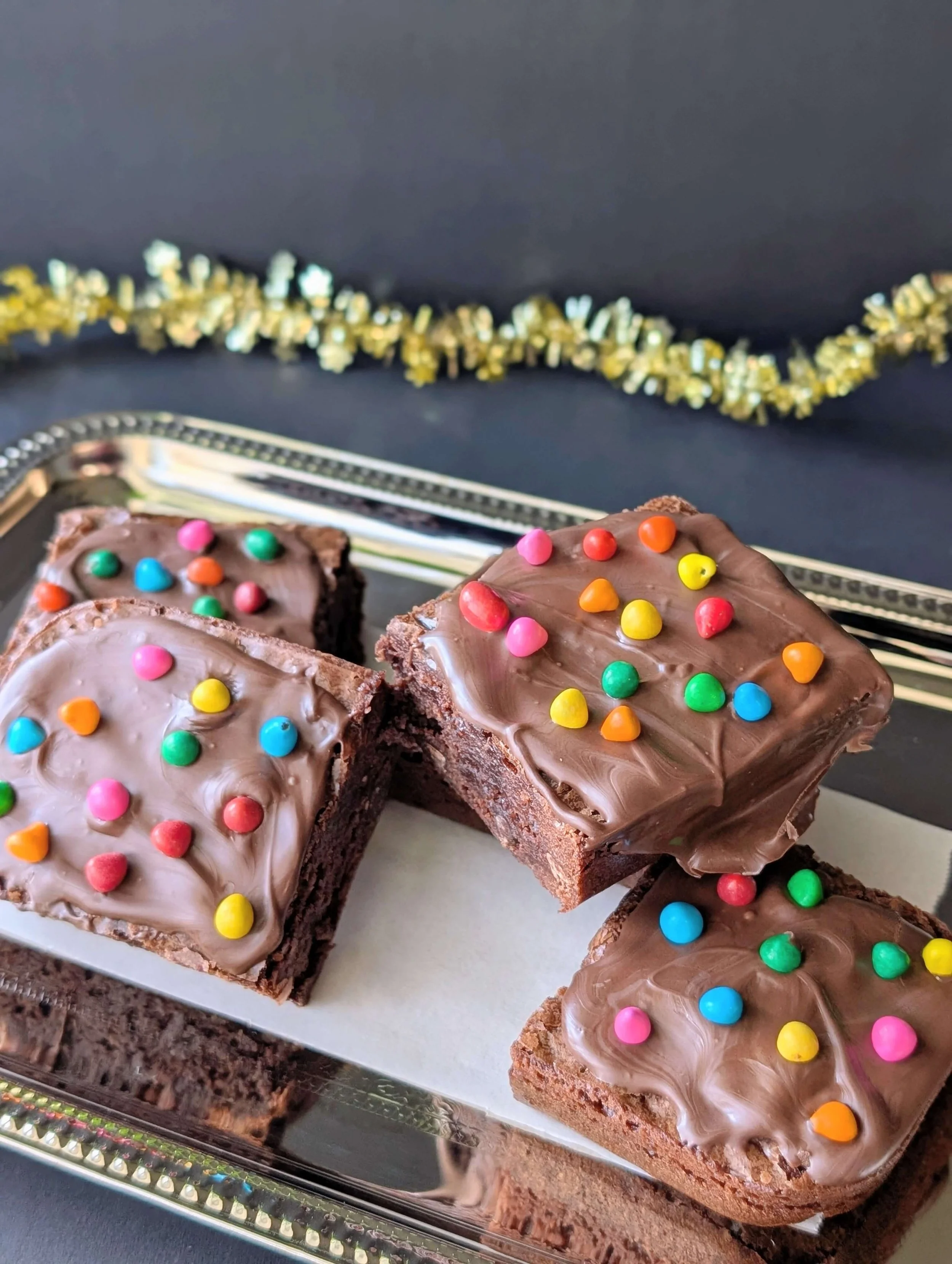 Four square, thick chocolate cosmic brownies topped with glossy milk chocolate frosting and colorful, candy-coated chocolate pieces, arranged on a silver serving platter with a blurred gold tinsel garland in the background.