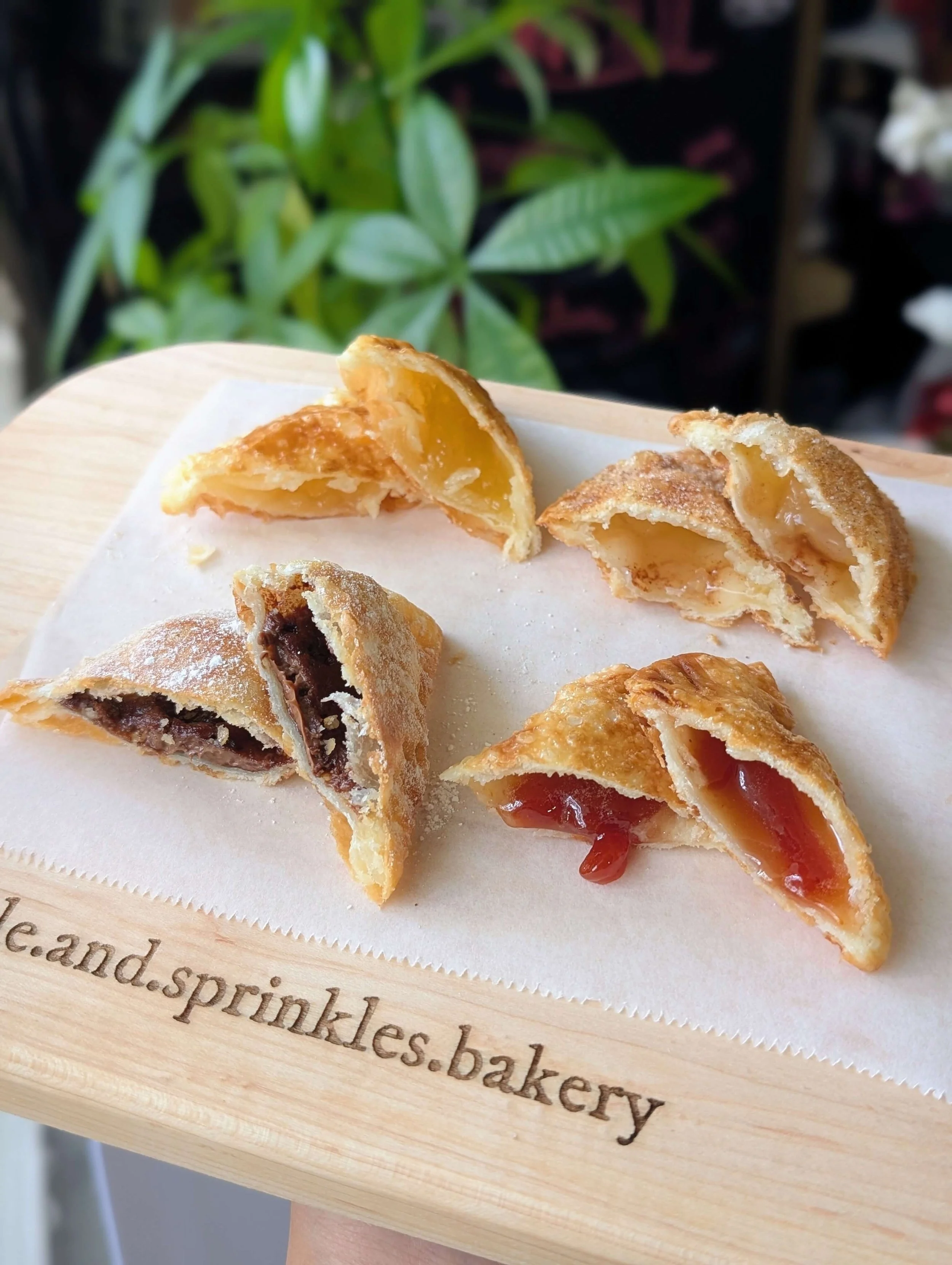Four sweet empanadas cut in half to showcase a variety of fillings—including Nutella dusted with powdered sugar, guava, apple pie, and pineapple—arranged on parchment paper atop a wooden board engraved with "@twinkle.and.sprinkles.bakery", with a blu