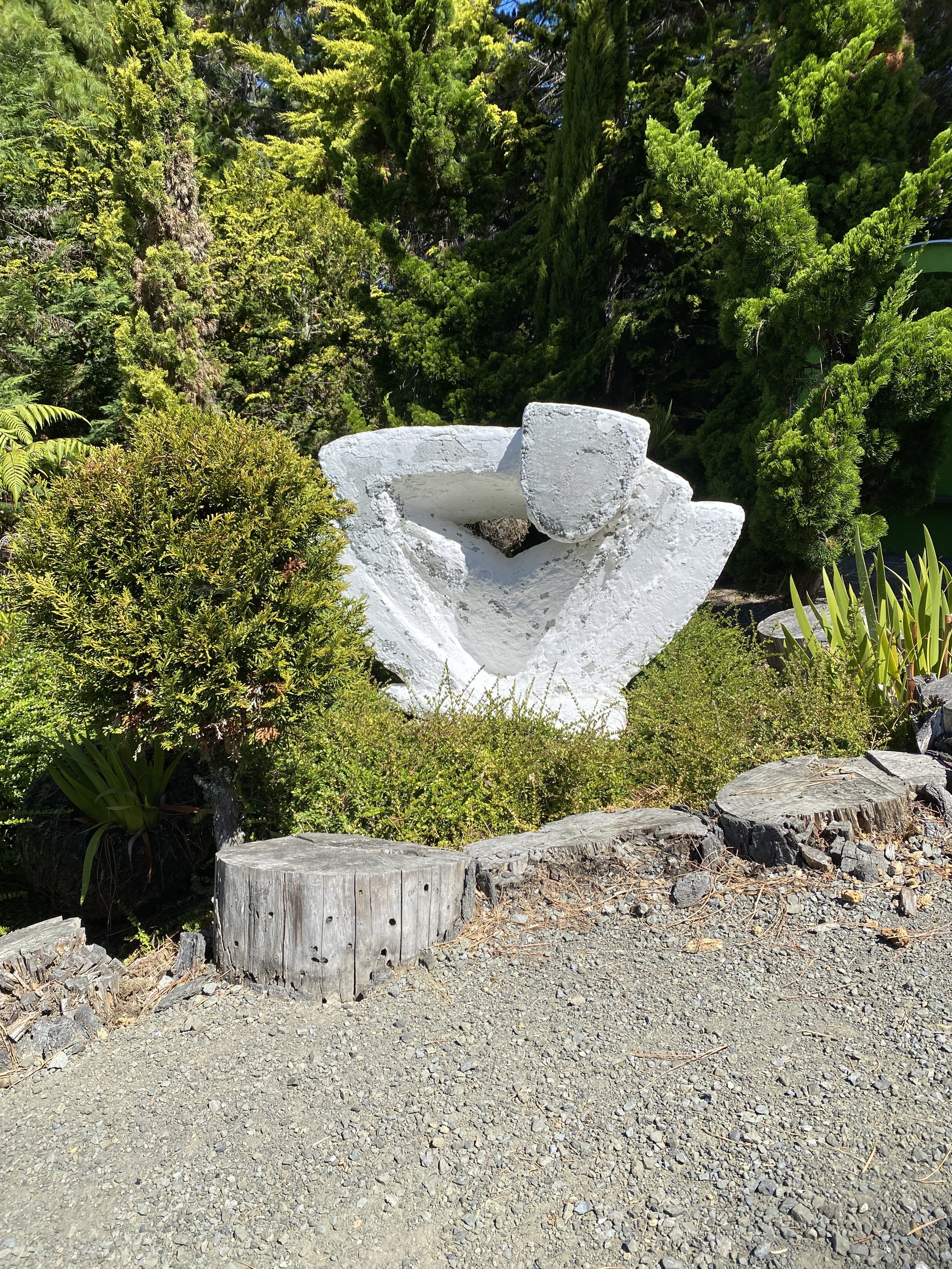 Abstract white sculpture in a garden surrounded by green foliage and tree stumps on gravel.
