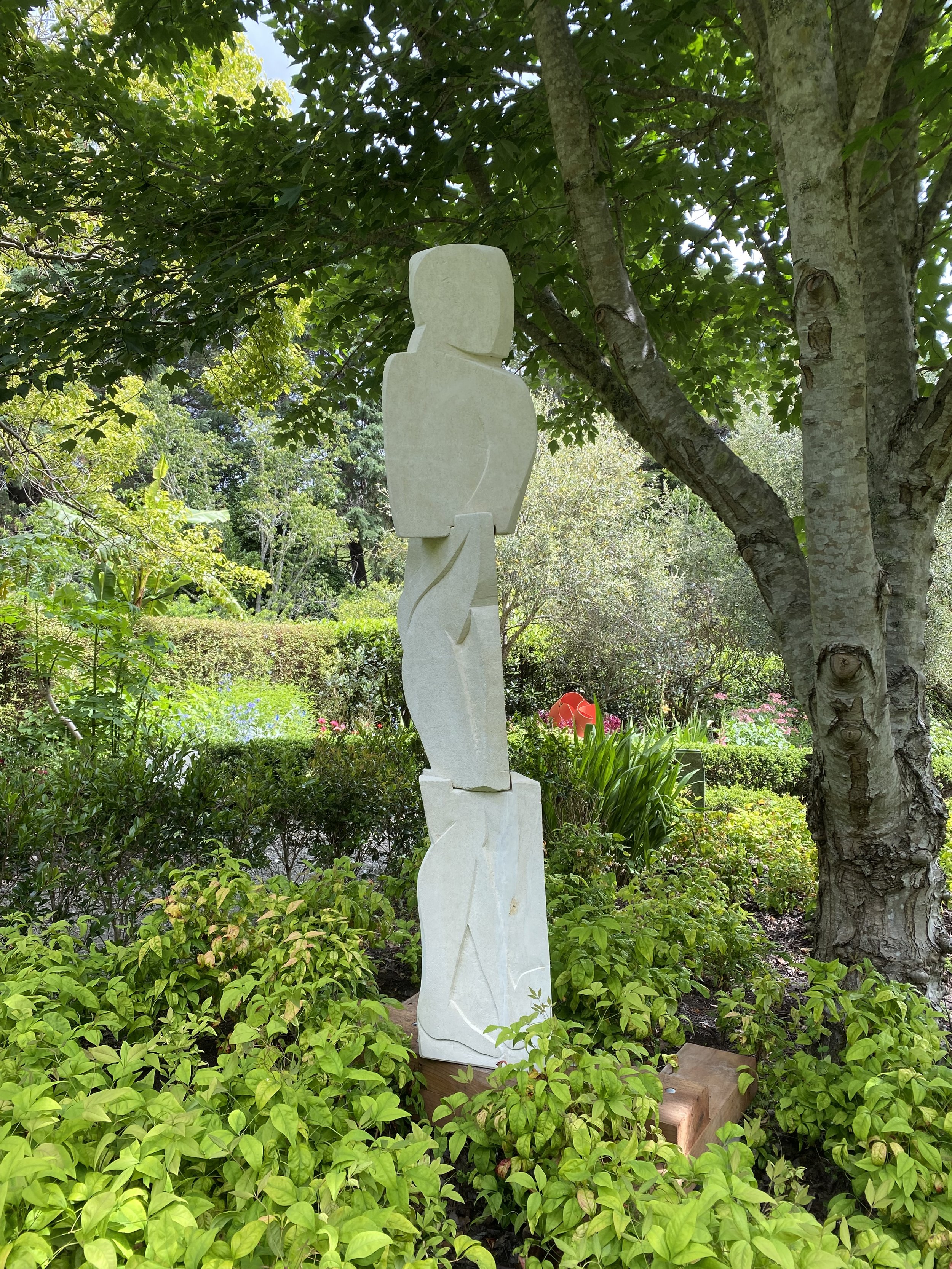 Abstract stone sculpture in a garden setting surrounded by green foliage and trees.