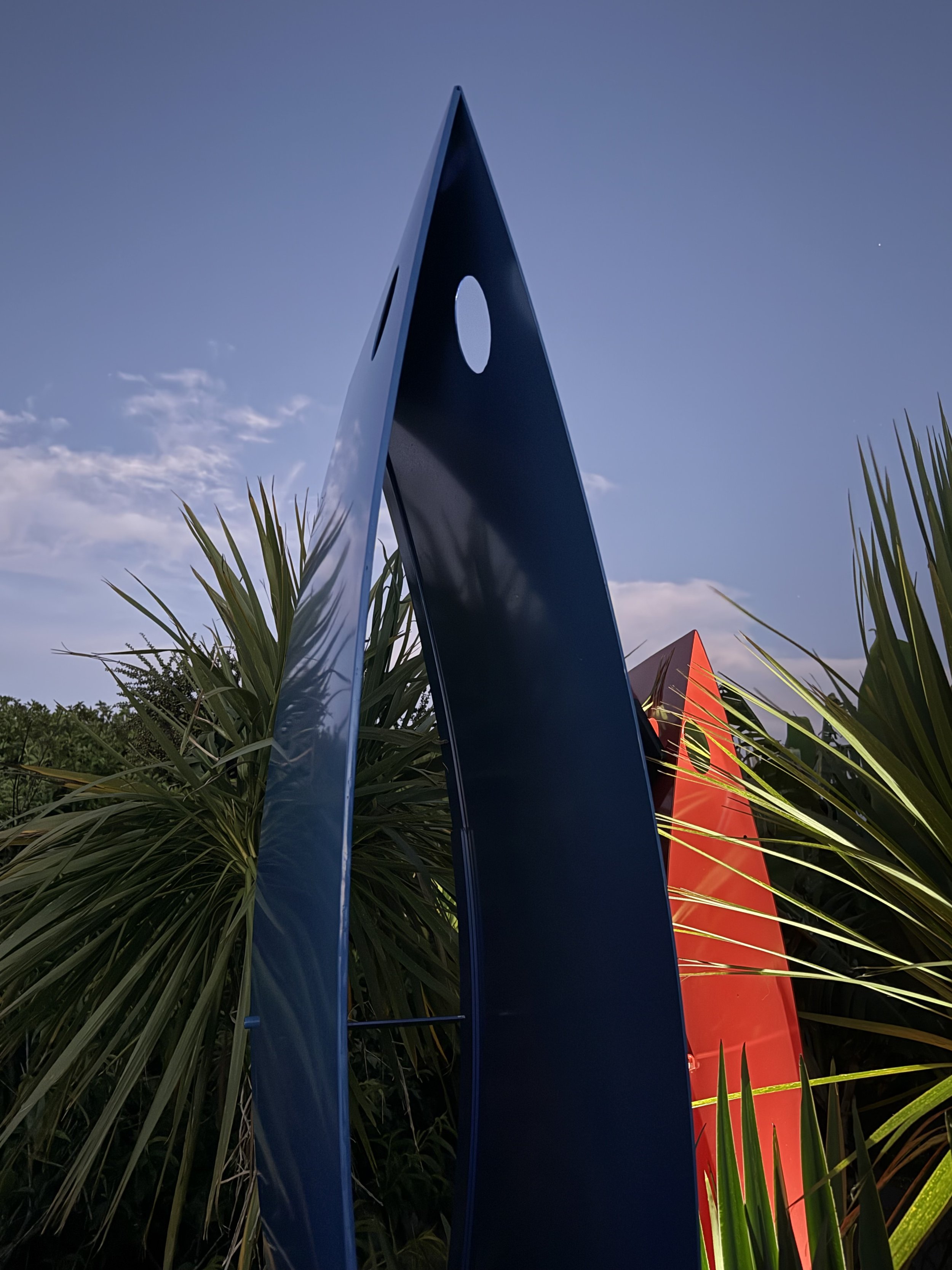 Colorful metal sculptures resembling tall, angular shapes with holes, surrounded by palm trees, under a clear blue sky.