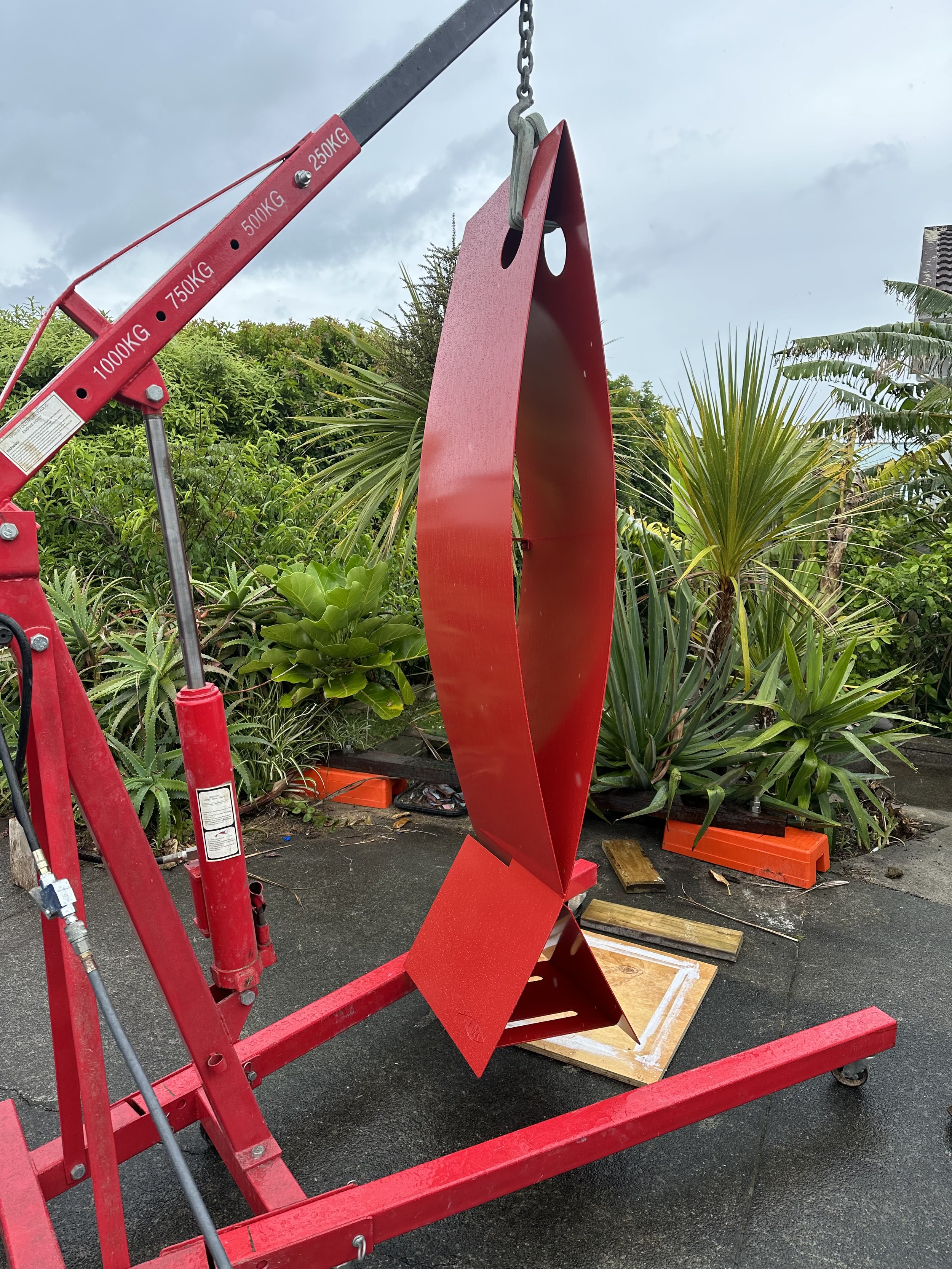 A red metal sculpture or object hanging from a crane in an outdoor setting with green plants in the background.