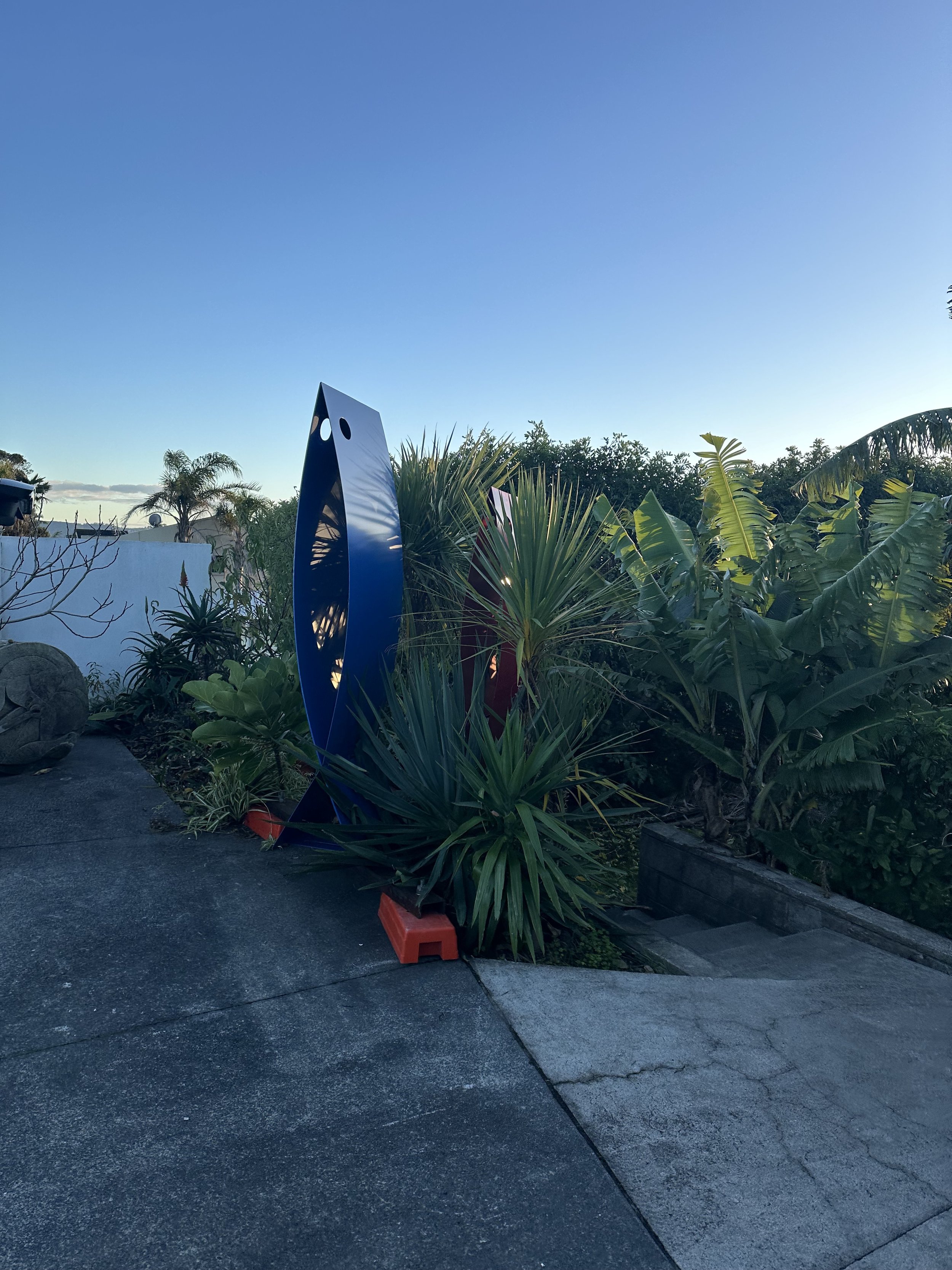 Outdoor scene with modern art sculptures, tropical plants, and a concrete pathway. The sculptures are blue and red, with sleek, abstract shapes. The area is landscaped with various green plants and there is a clear blue sky above.