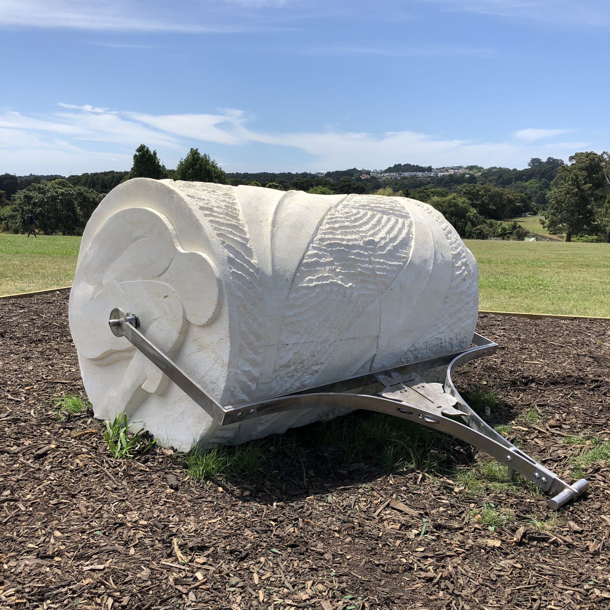Abstract stone sculpture resembling a rolled-up scroll with intricate carvings, mounted on metal supports, set in a park landscape.