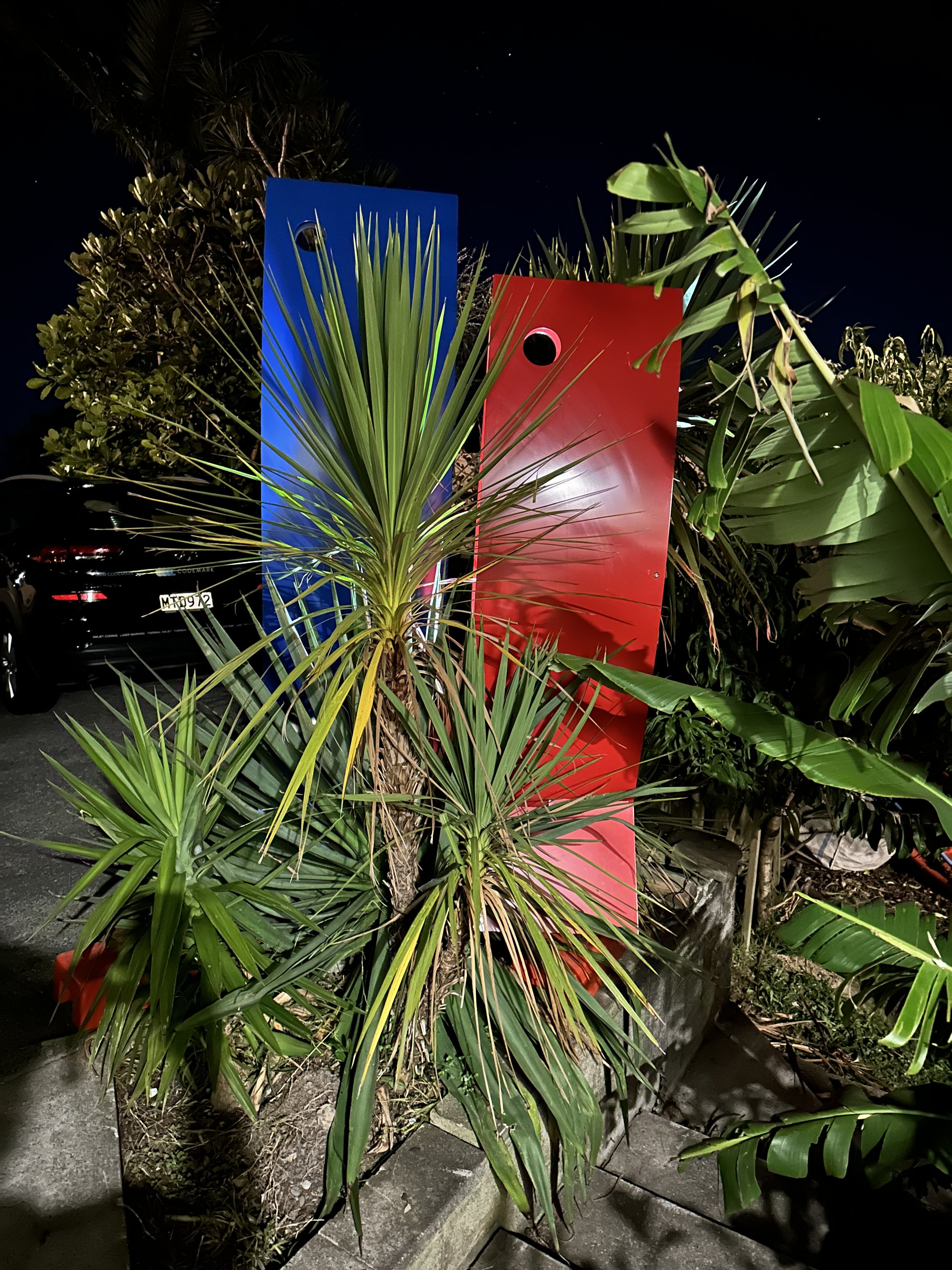 Outdoor scene with green tropical plants and sculptures at night, featuring red and blue metal panels behind foliage and a parked car in the background.