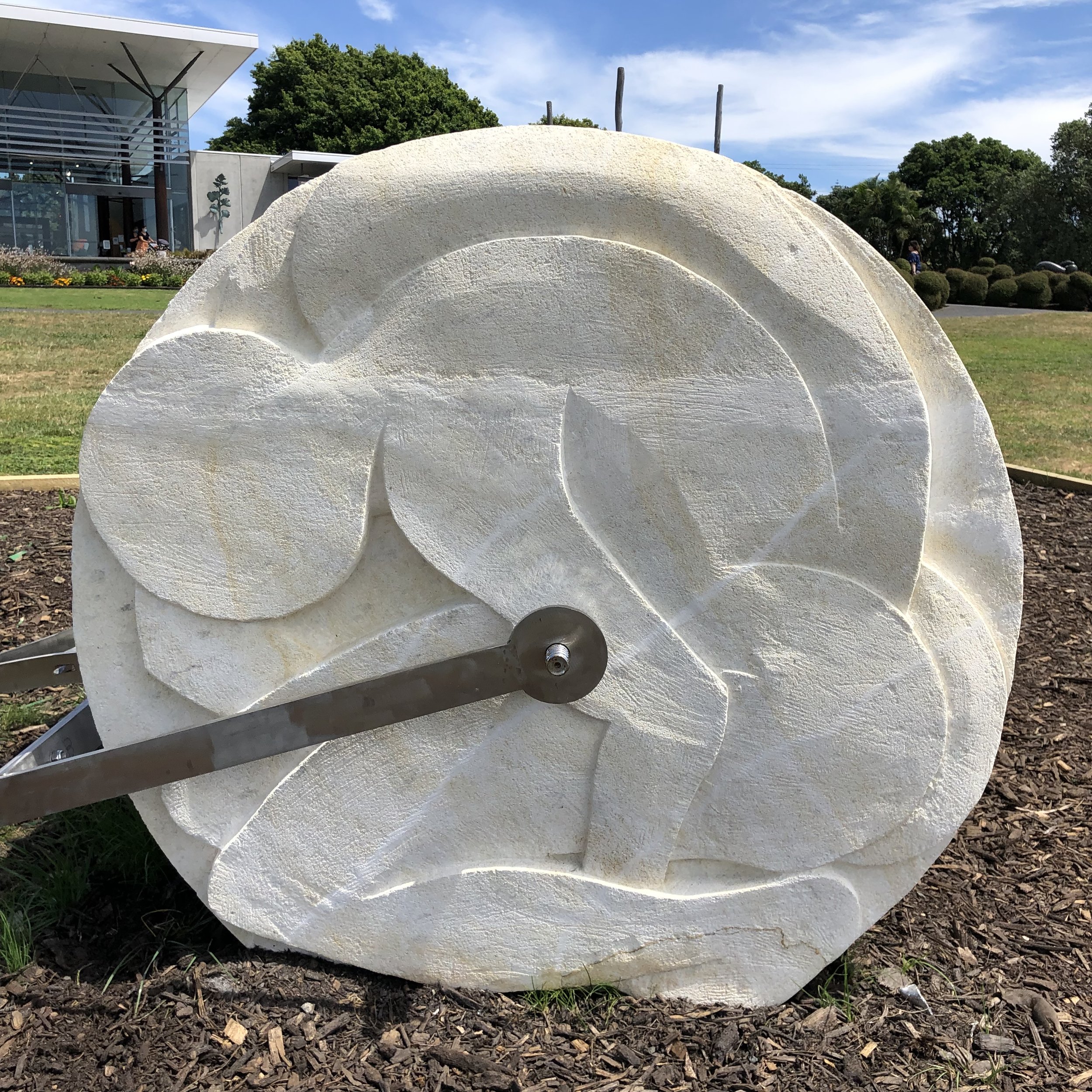 Large stone wheel with abstract carvings, featuring attached metal lever, situated in a garden setting with building and trees in the background.