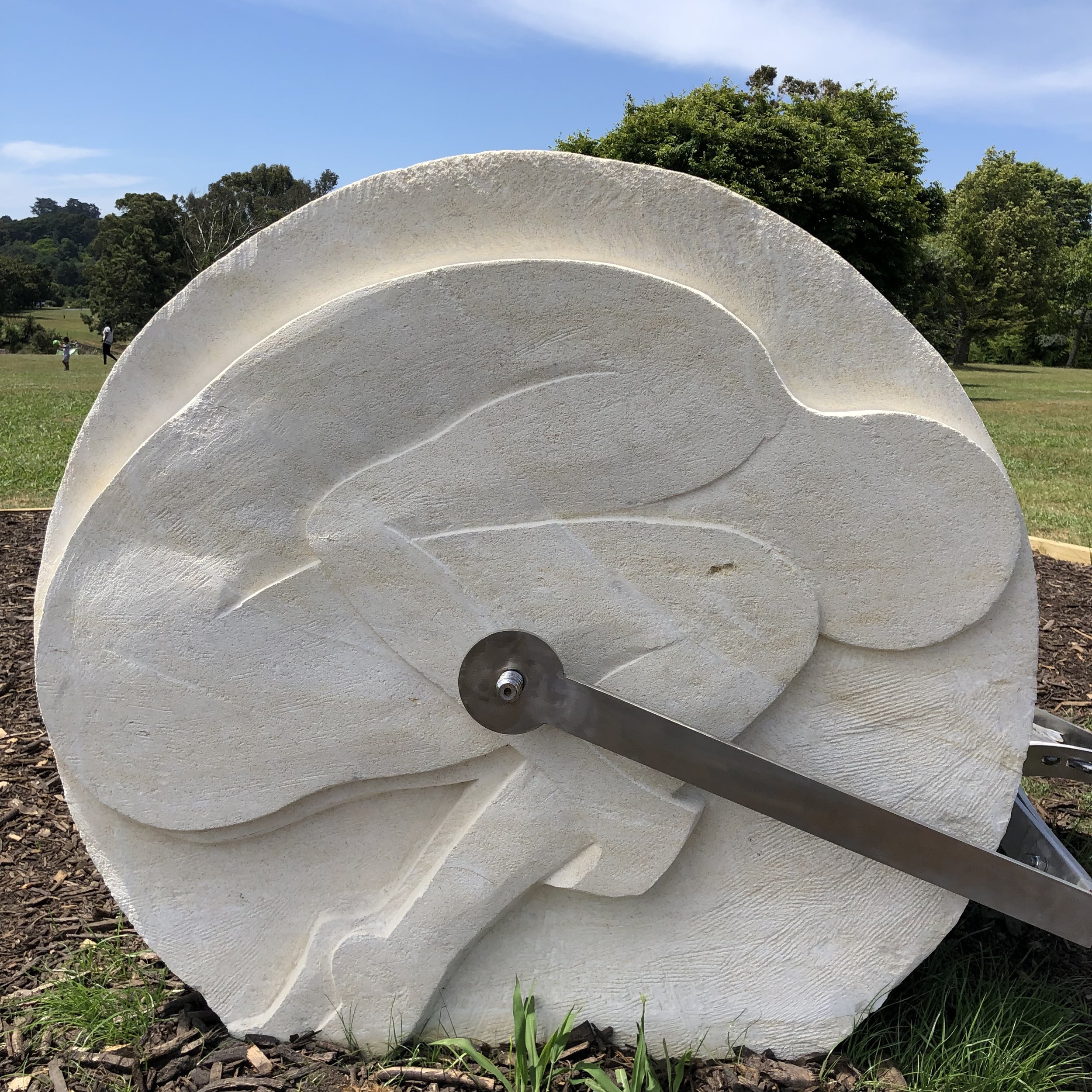 White stone sculpture depicting a crouched human figure embedded in a circular shape, outdoors with grass and trees in the background.
