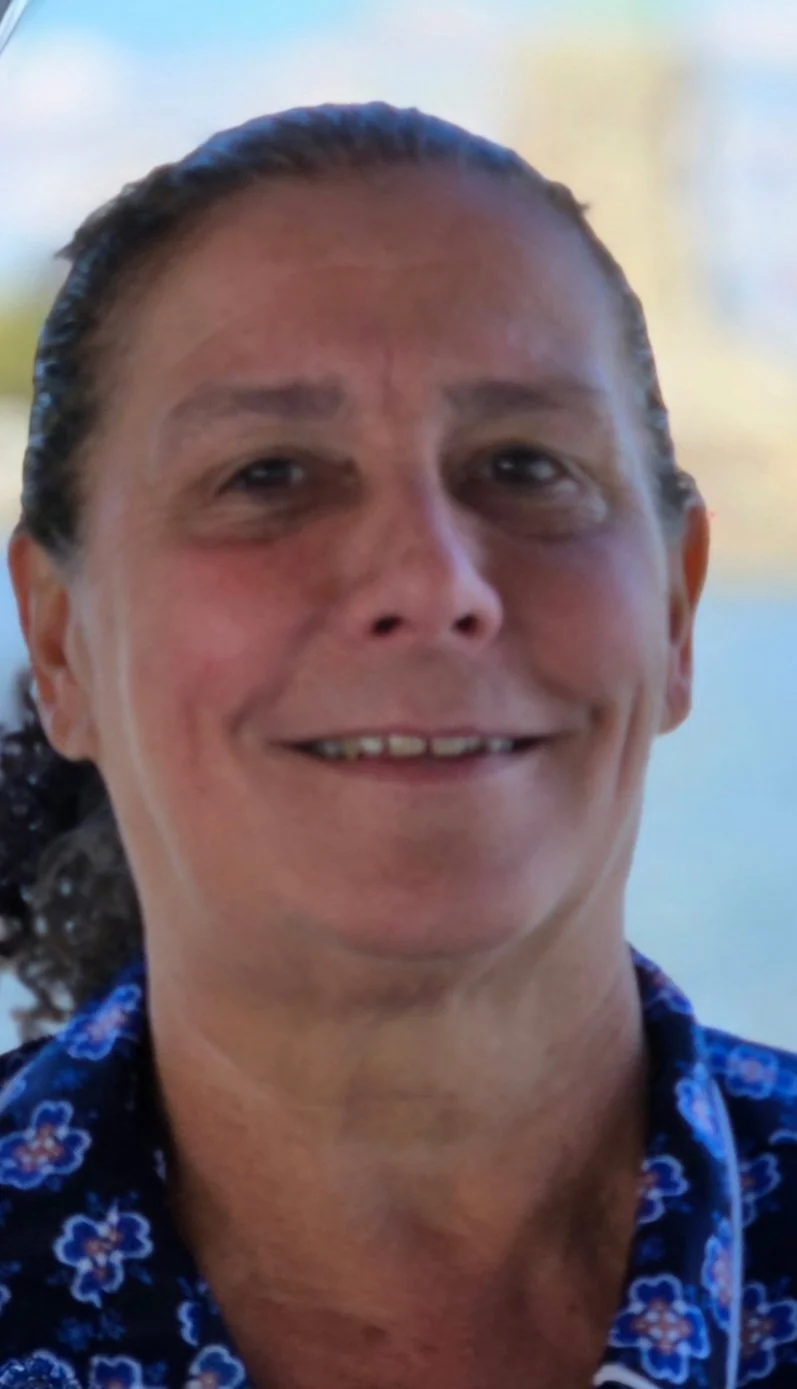 Close-up of a smiling woman with dark hair pulled back, wearing a blue floral shirt, outdoors with blurry background.