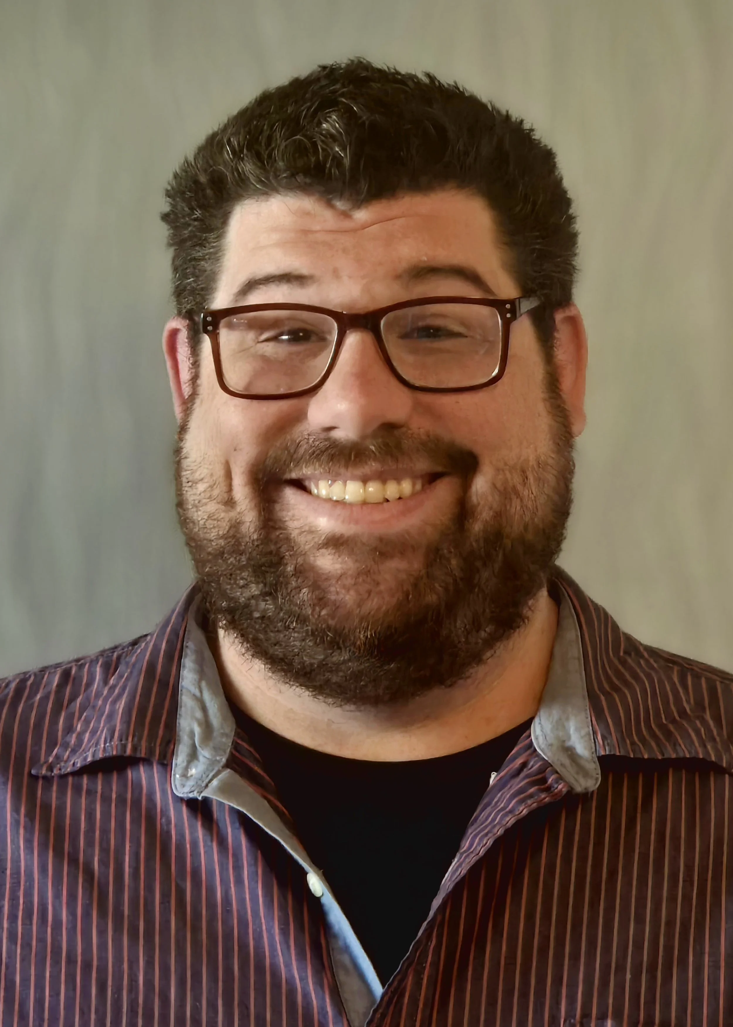 A man with glasses and a beard smiling, wearing a striped collared shirt.