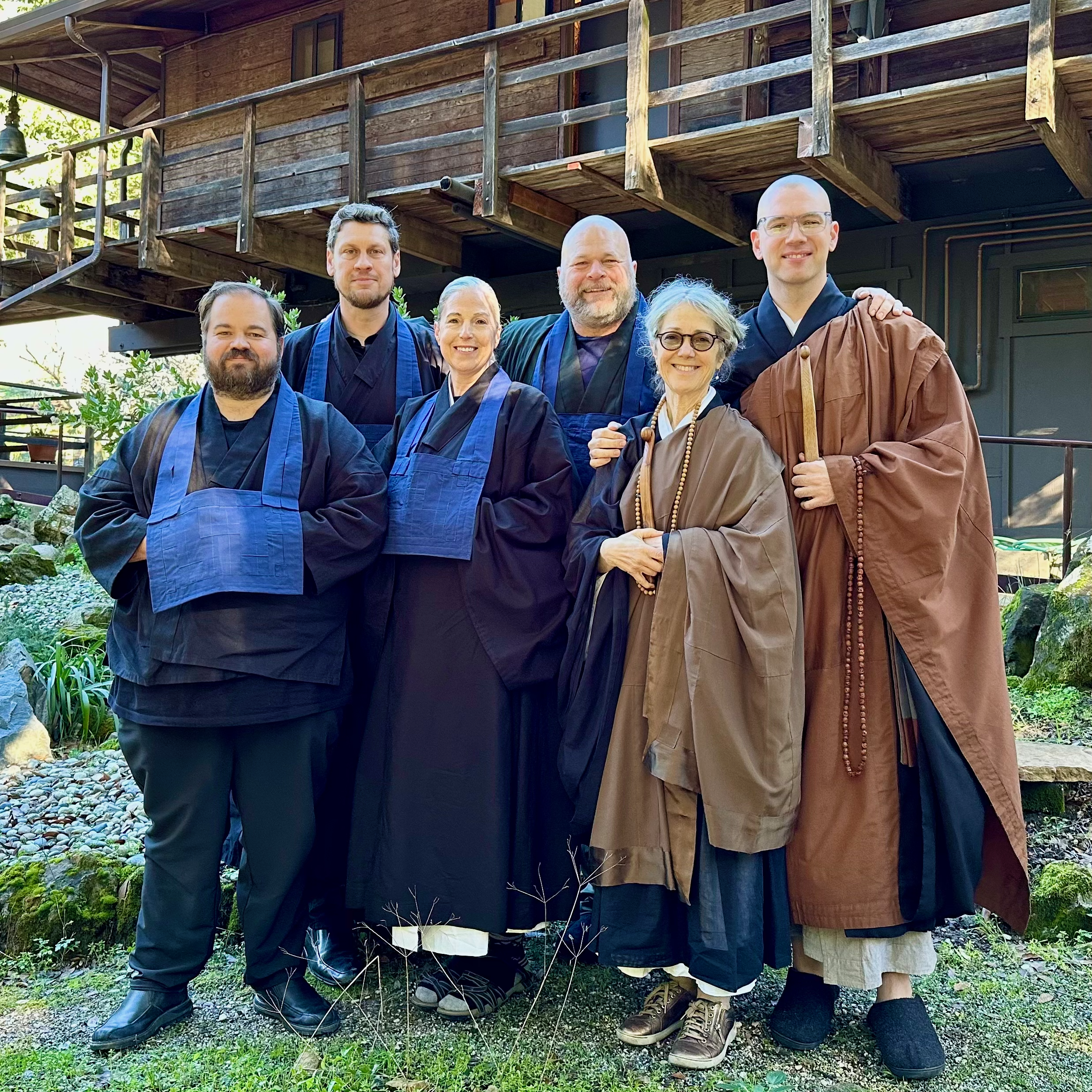 A group of six people in robes outside at Jikoji Zen Center. Thor Keika Cary, Matt Unzan Meinhardt, Öshin Jennings Sensei, Jesse Rentai Parrish Lewis, Tam Getsuki Schmidt, Pamela Nenzen Brown Sensei. All are smiling.