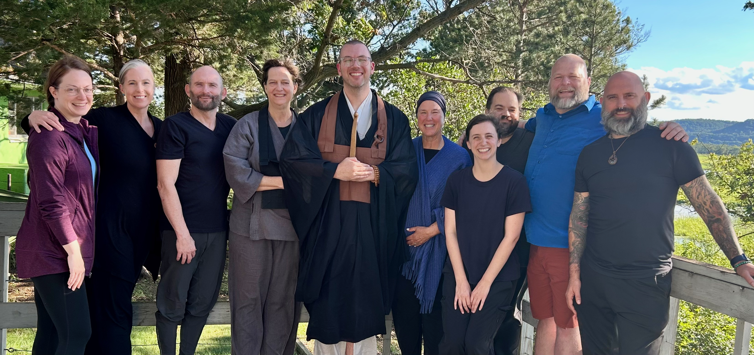 A group of ten smiling people standing outside after the NBZ Minnesota Sesshin in August 2023. Behind them is lush green foliage and a river flowing.