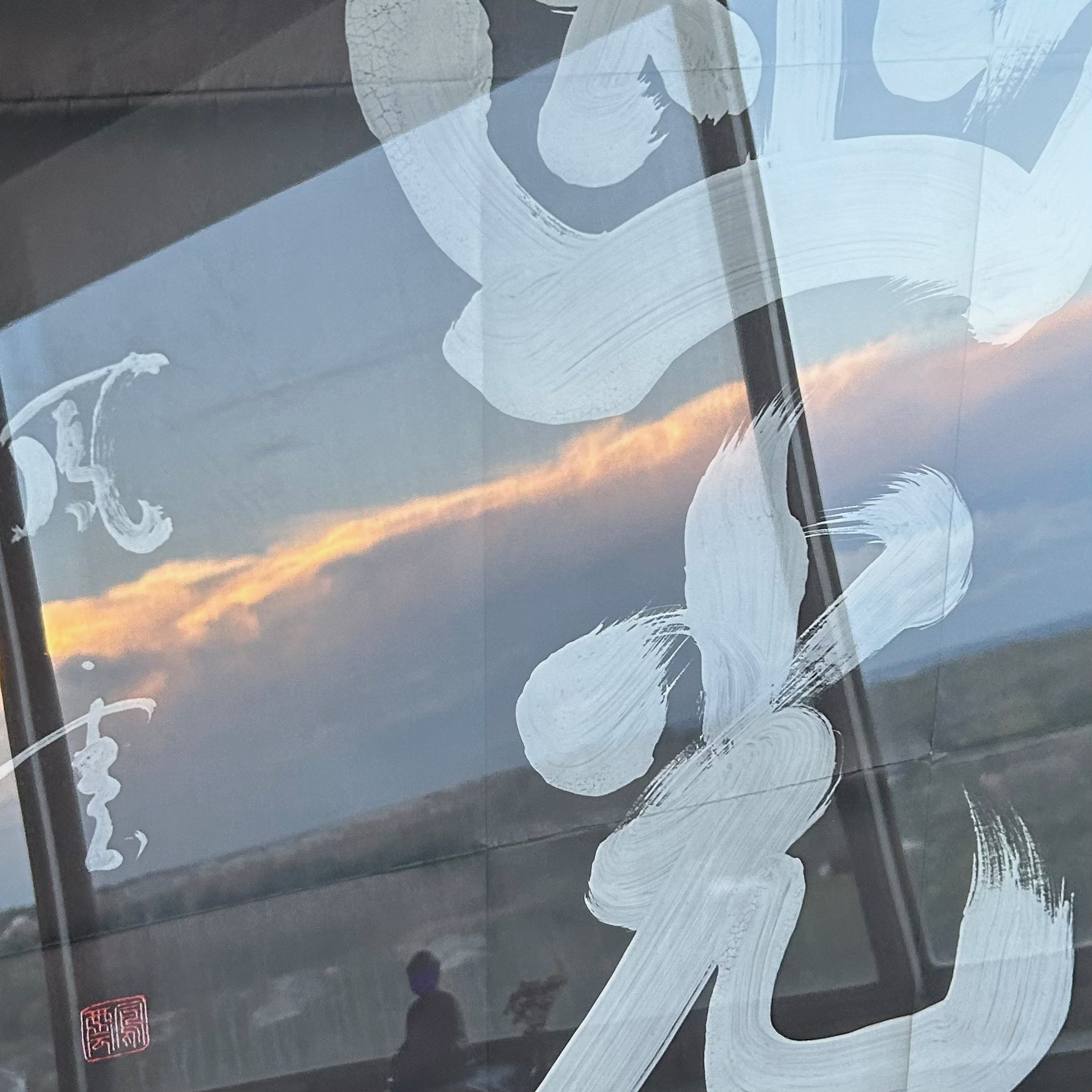 A large print of white kanji "Bodhi" on a black background. In the reflection found on the print's front, there is a brilliant sunset across the fluffy clouds, and a small Buddha statue on the window sill is reflected over Maryland.