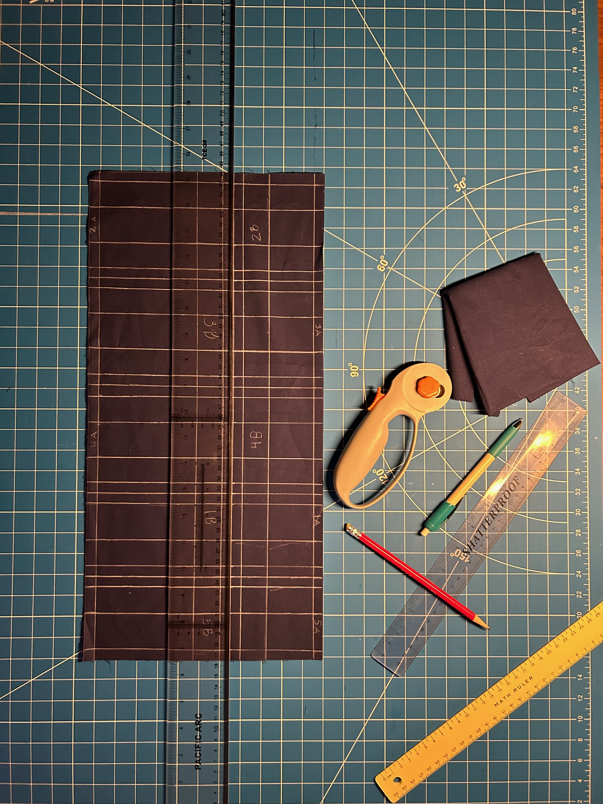 A collection of sewing tools are scattered neatly on a sewing mat next to a piece of blue rectangular fabric that is covered in chalk lines and markings, destined to be a rakusu.