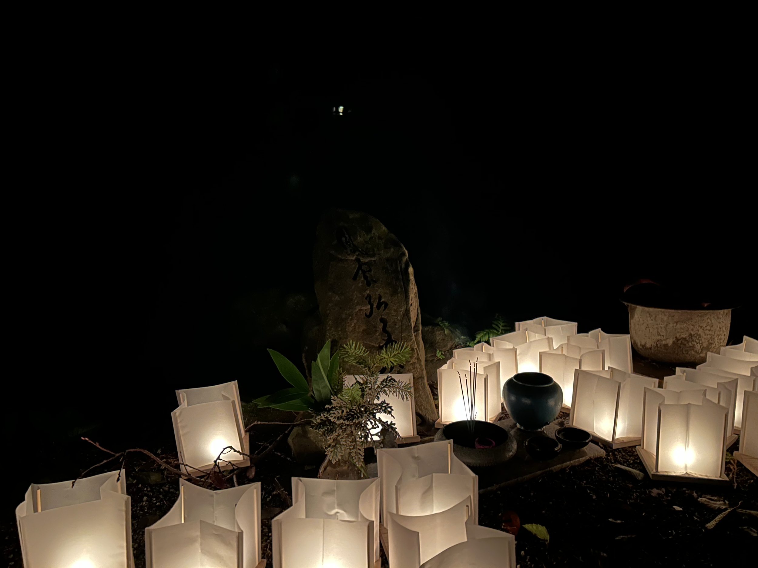 A group of paper lanterns lights up a stone memorial tablet with carved kanji in the dark of night.