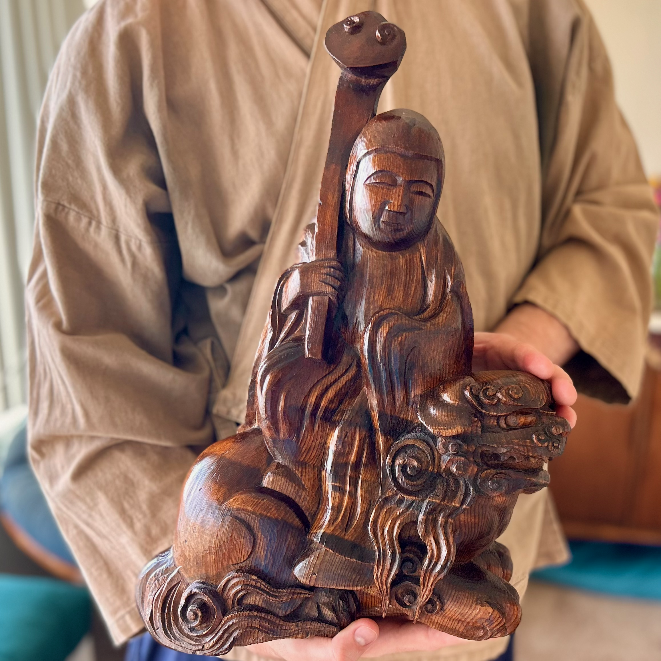 A brown mottled wooden statue on Manjuśri, the Bodhisattva of Wisdom, riding a formidable lion, whose mane transforms into clouds around the base. Manjuśri carries a lotus staff and has a soft, serene expression. The statue is held by a white man wea
