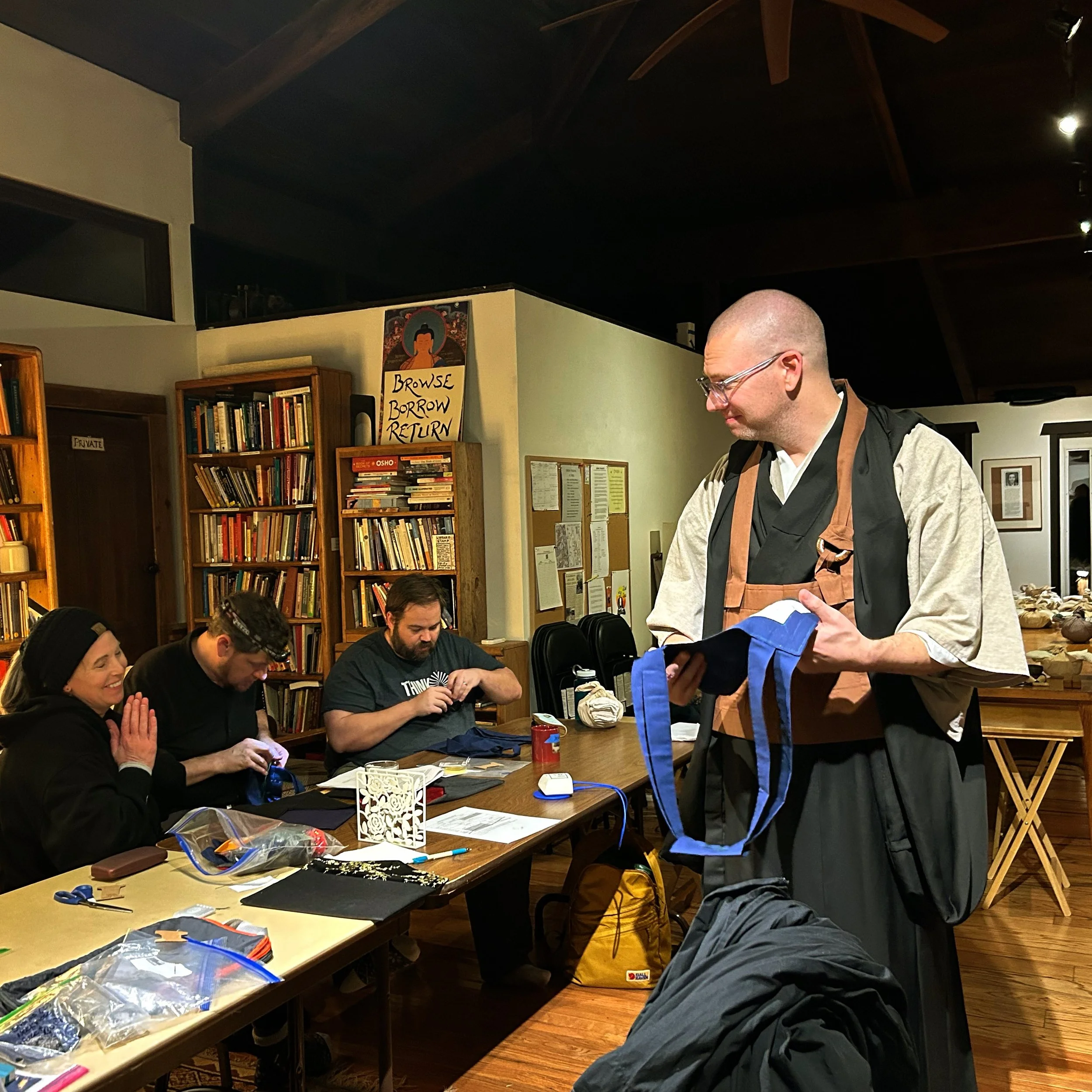Ōshin Sensei, Tam Getsuki, Thor Keika, and Jesse Rentai are gathered at a table sewing their rakusu with several full bookshelves behind them. Ōshin tenderly holds a Tam's rakusu while smiling at her.