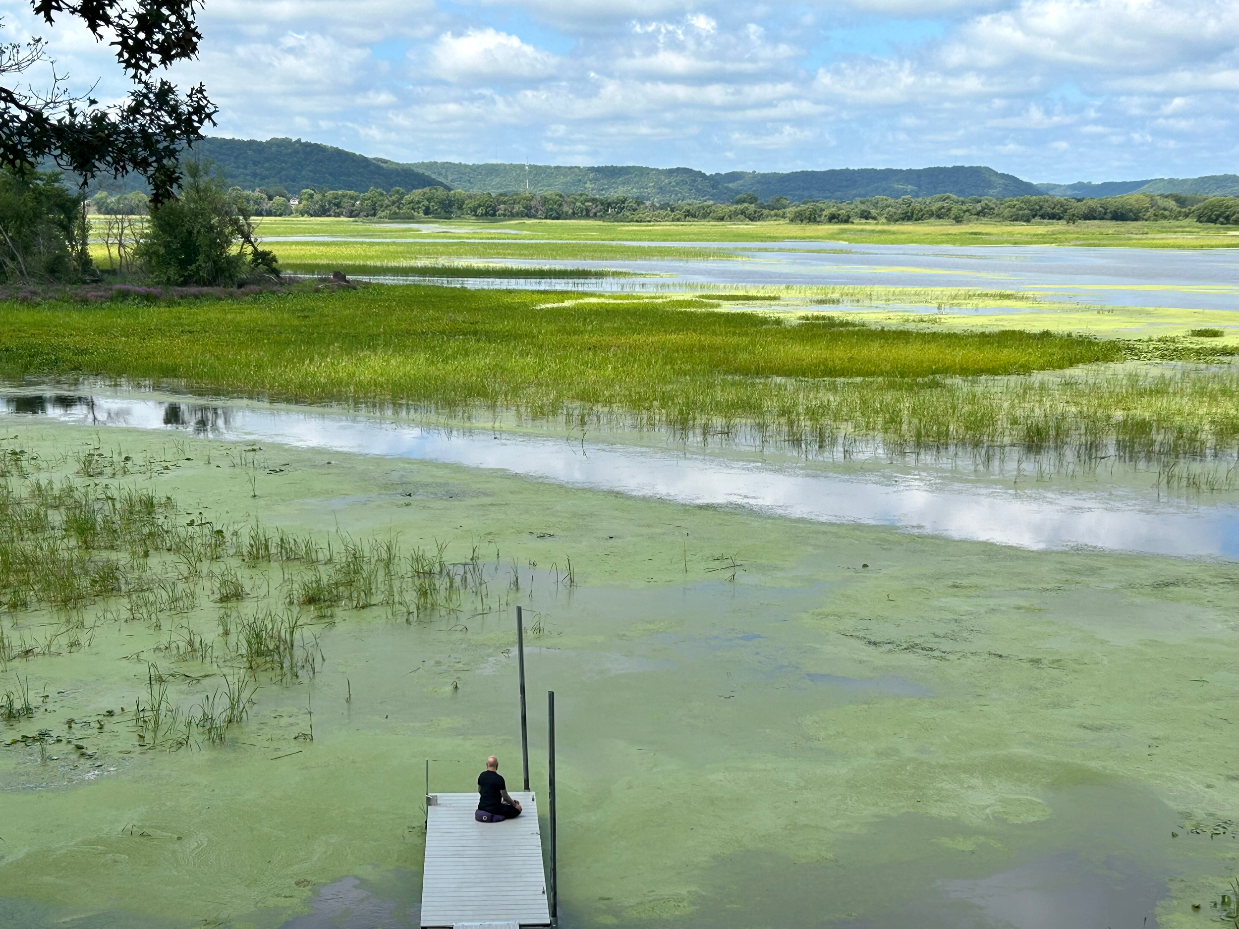 Swampy land meets a wide flowing river in northern Minnesota. In the distance, wide hills rise up, reaching into the cloudy, pale blue sky. In the foreground, a dock extends into the water. There is someone meditating at the very end of the dock.