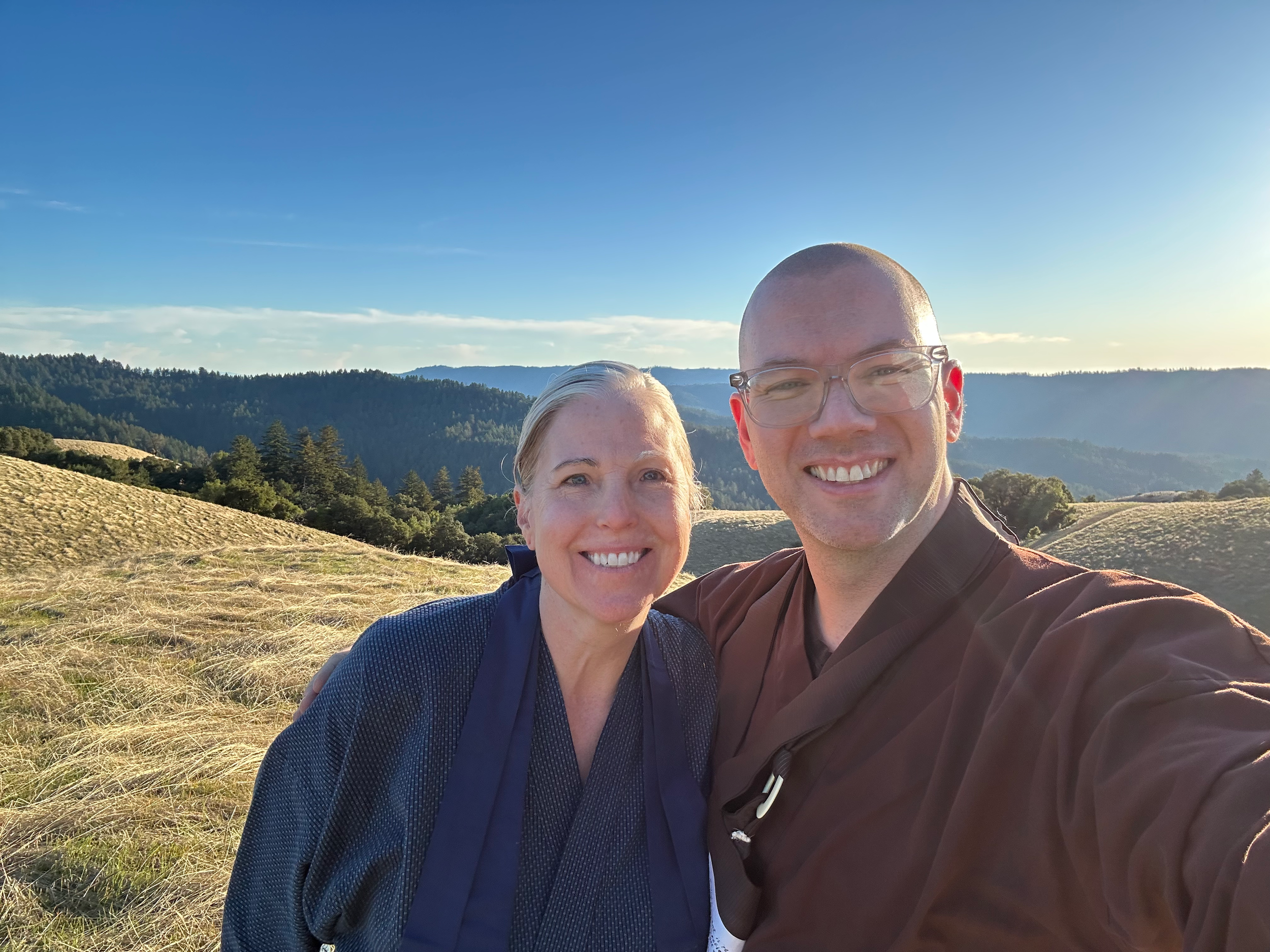 Getsuki, a white woman with a warm smile and white hair pulled back in a bun, is standing next to Ōshin Sensei, a white man with clear classes and a friendly smile. They are at the Ridge in Jikoji, the top of a series of hills covered in grasses and