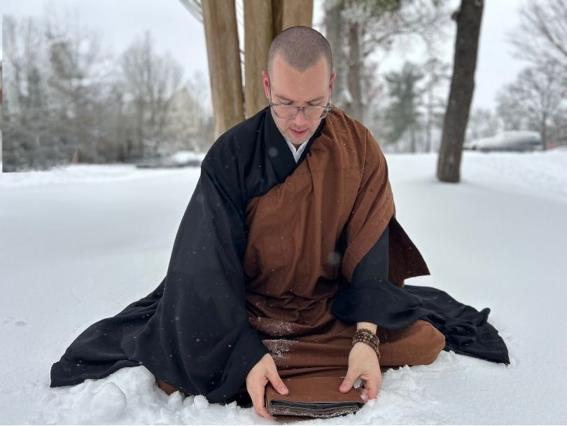 Ōshin, a white man with a shaved head, is sitting in the snow in front of a tree. He is wearing Buddhist robes, surrounded by snow-covered trees in the background. He is carefully placing his folded zagu in front of him, preparing to begin zazen.