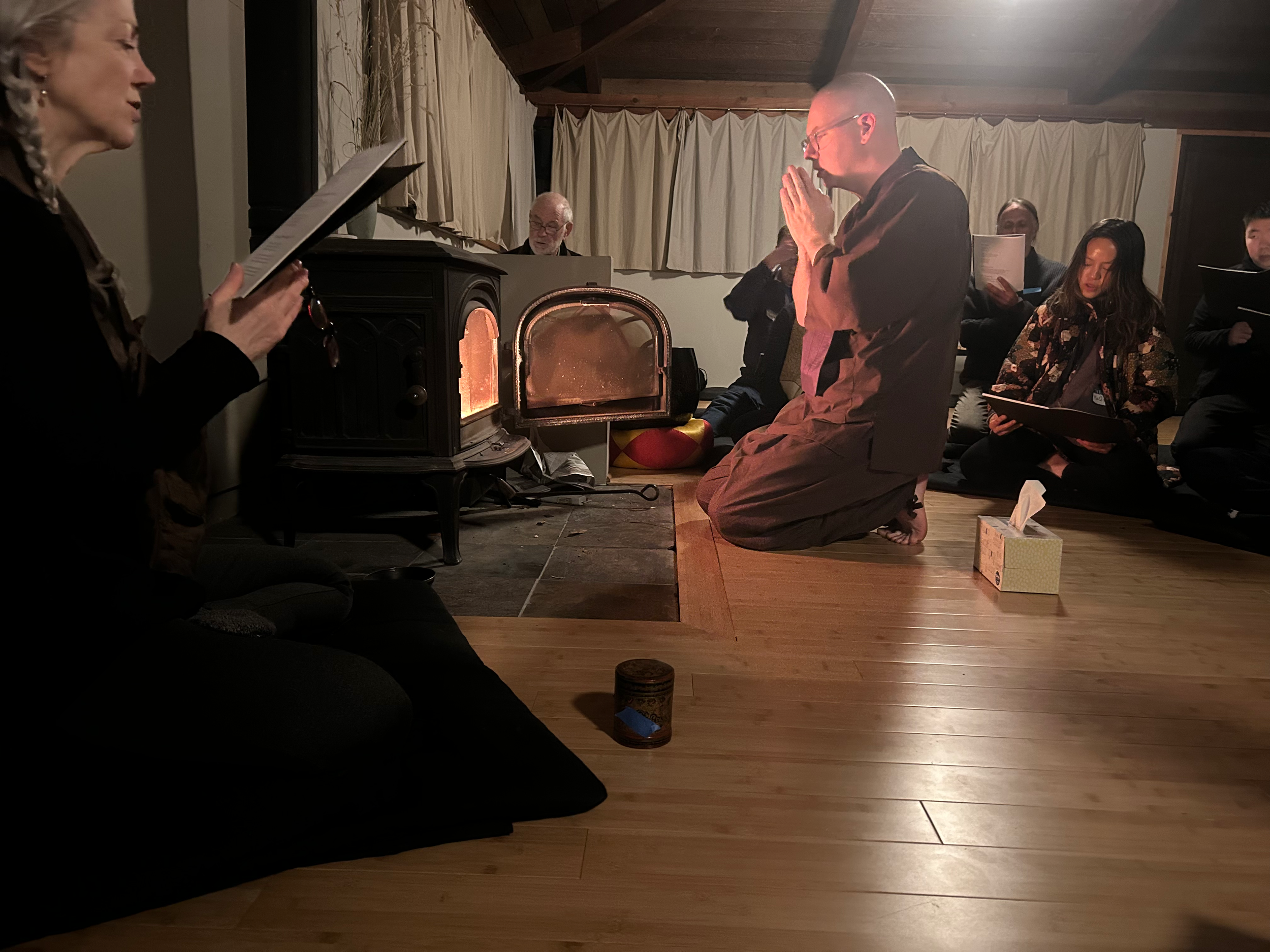 Ōshin Sensei, wearing brown samu-e and a brown rakusu, is kneeling in front of the large wood burning stove in Jikoji's main Zendo. His hands are in gassho as he makes an offering. There are several people gathered around him, chanting and preparing