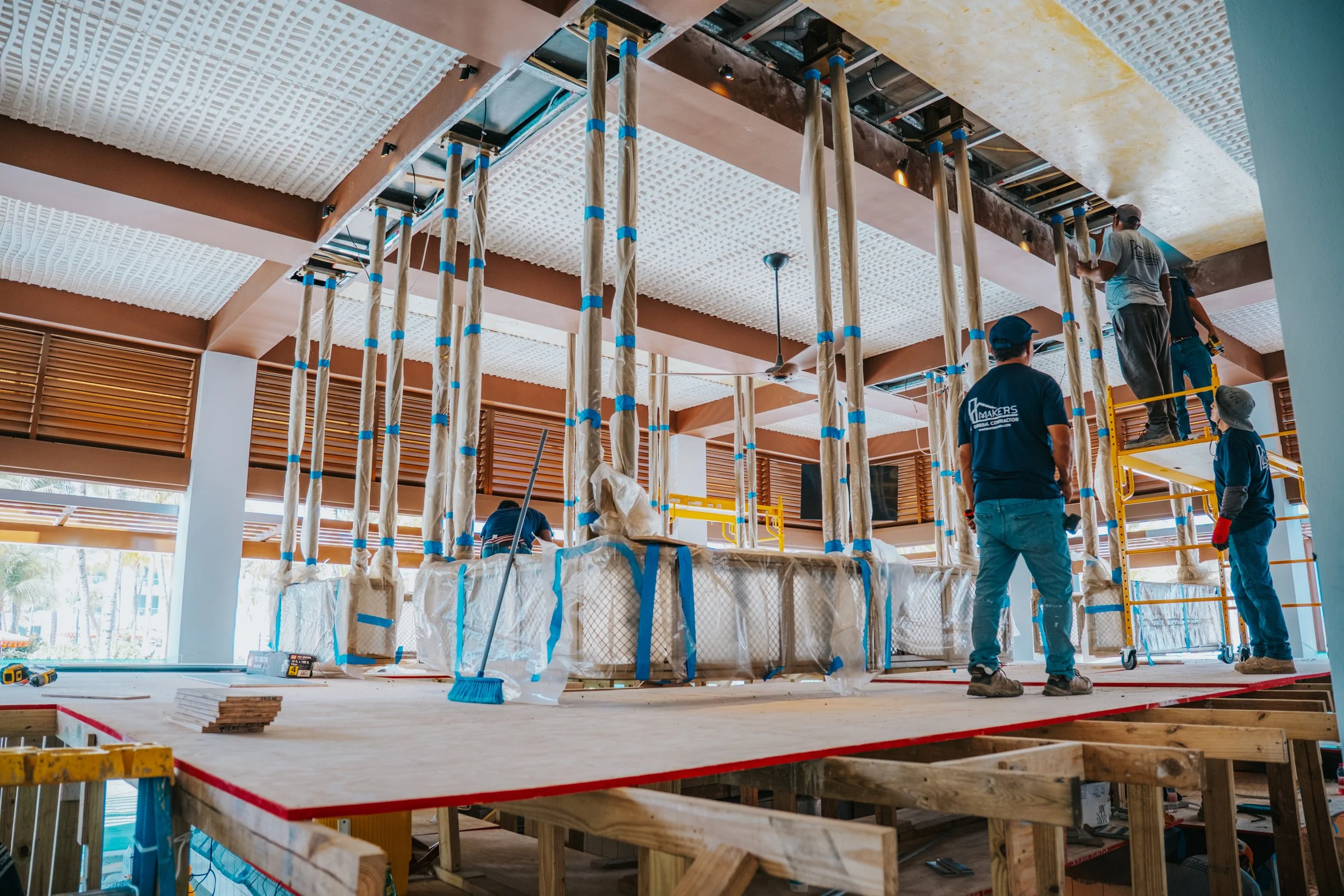 Construction workers installing ceiling panels in an unfinished indoor space with scaffolding and building materials.