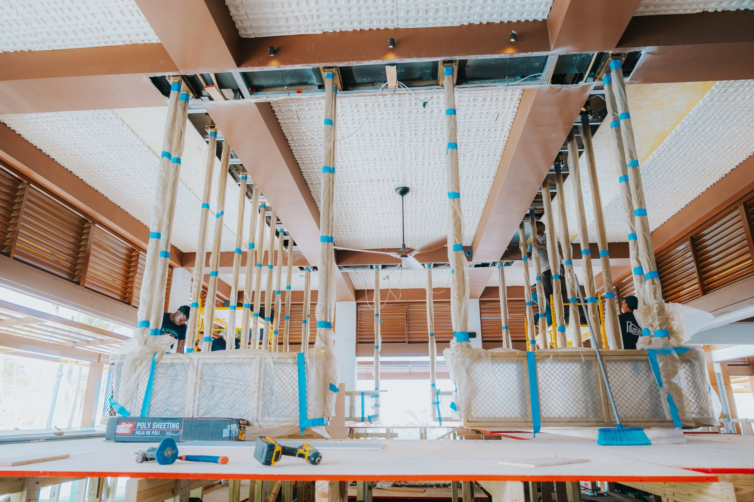 Construction workers on a scaffolding inside a building under renovation, installing drywall ceiling panels with tools and materials scattered on the floor.