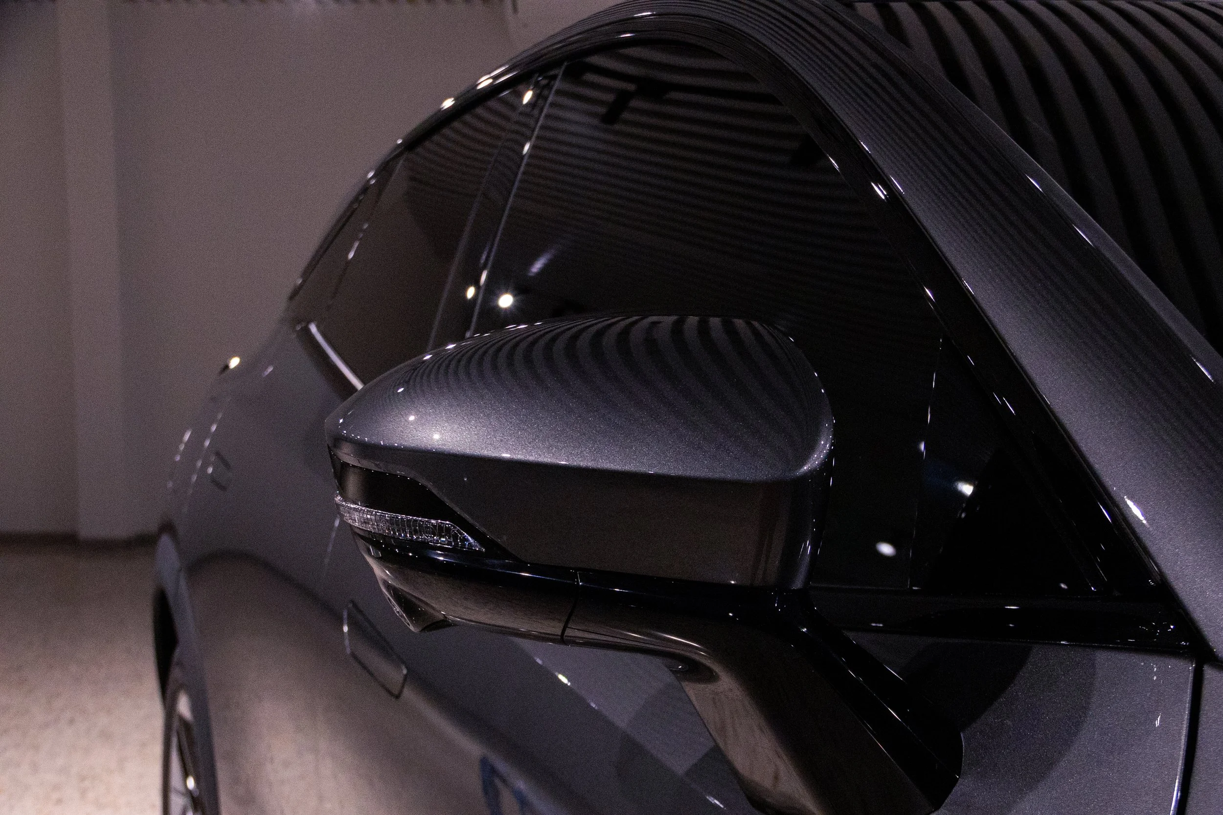 Close-up of a sleek, dark-colored car's side mirror with a patterned design, reflecting subtle light and surrounding environment.
