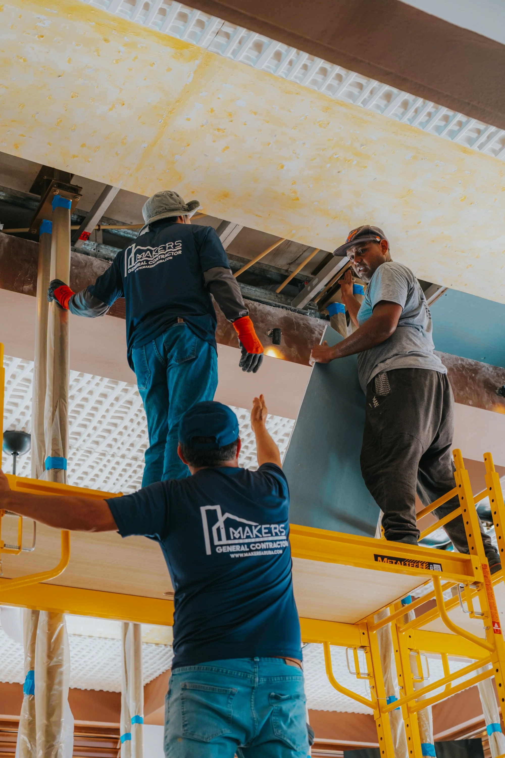 Two construction workers installing a ceiling panel, one standing on scaffolding and the other on the ground, both wearing work clothes and safety gear.