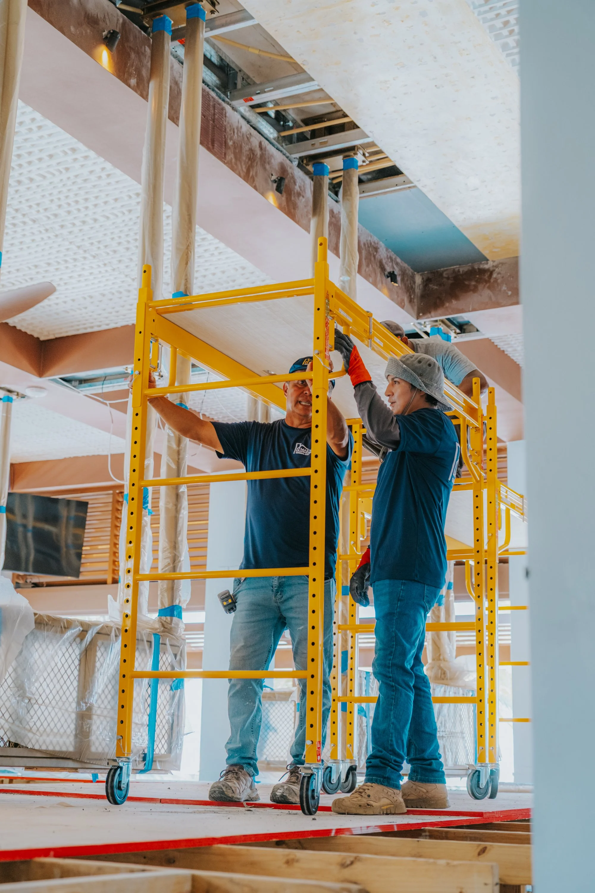 Construction workers installing or inspecting ceiling panels inside a building under construction, with scaffolding and construction materials visible.