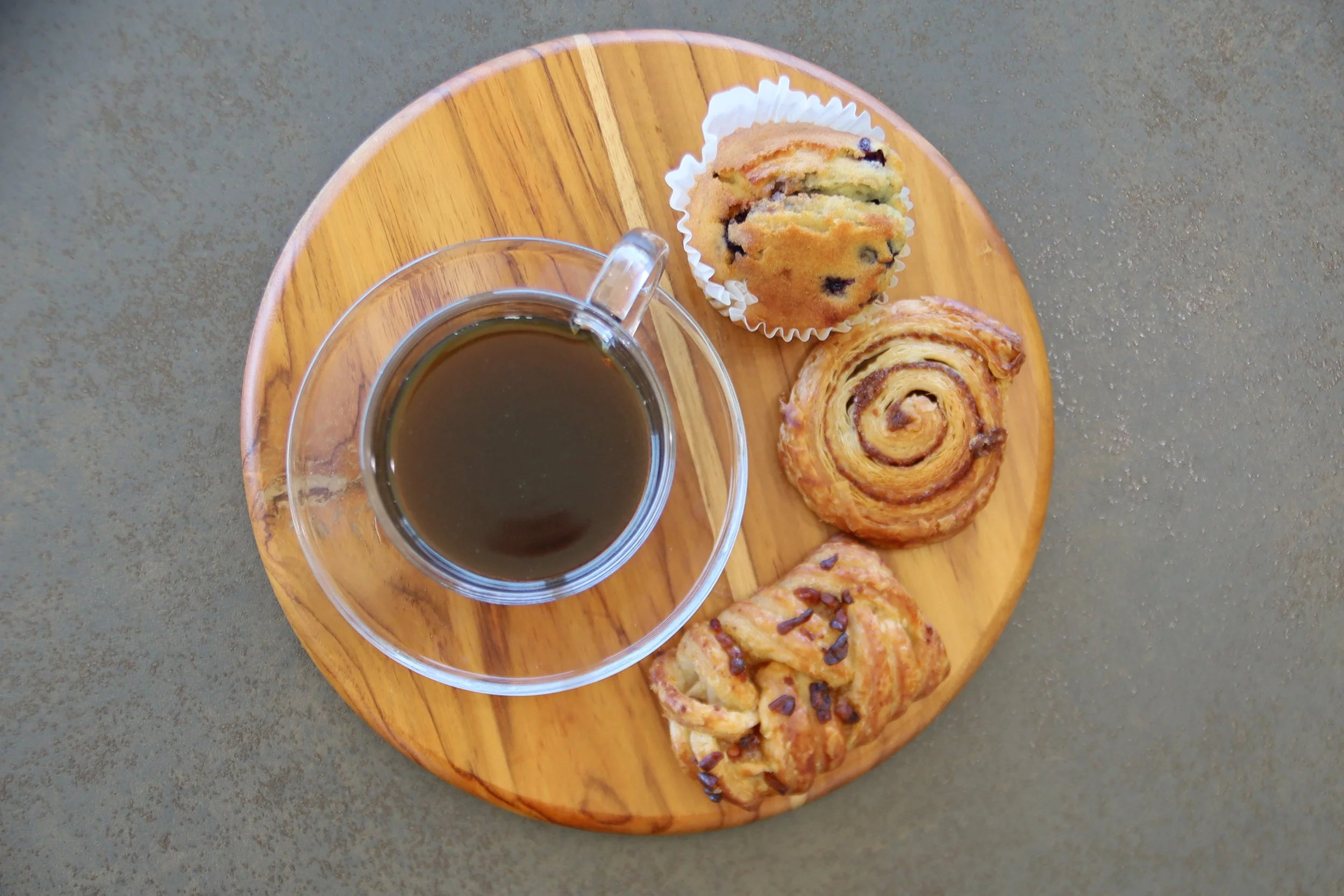 A glass cup of black coffee on a wooden board with a blueberry muffin, cinnamon roll, and pastry.