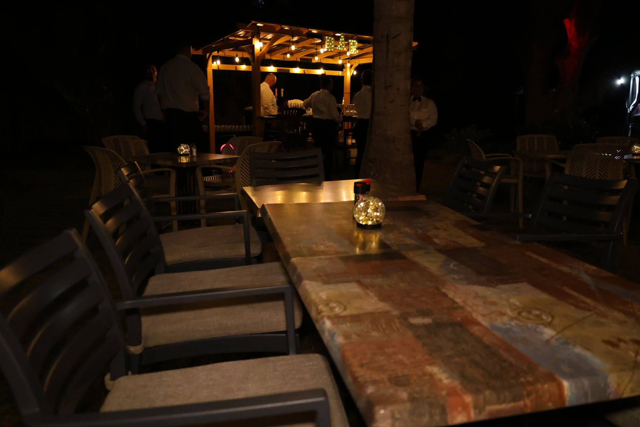Outdoor dining area at night with empty tables, chairs, and a decorated bar in the background with string lights.