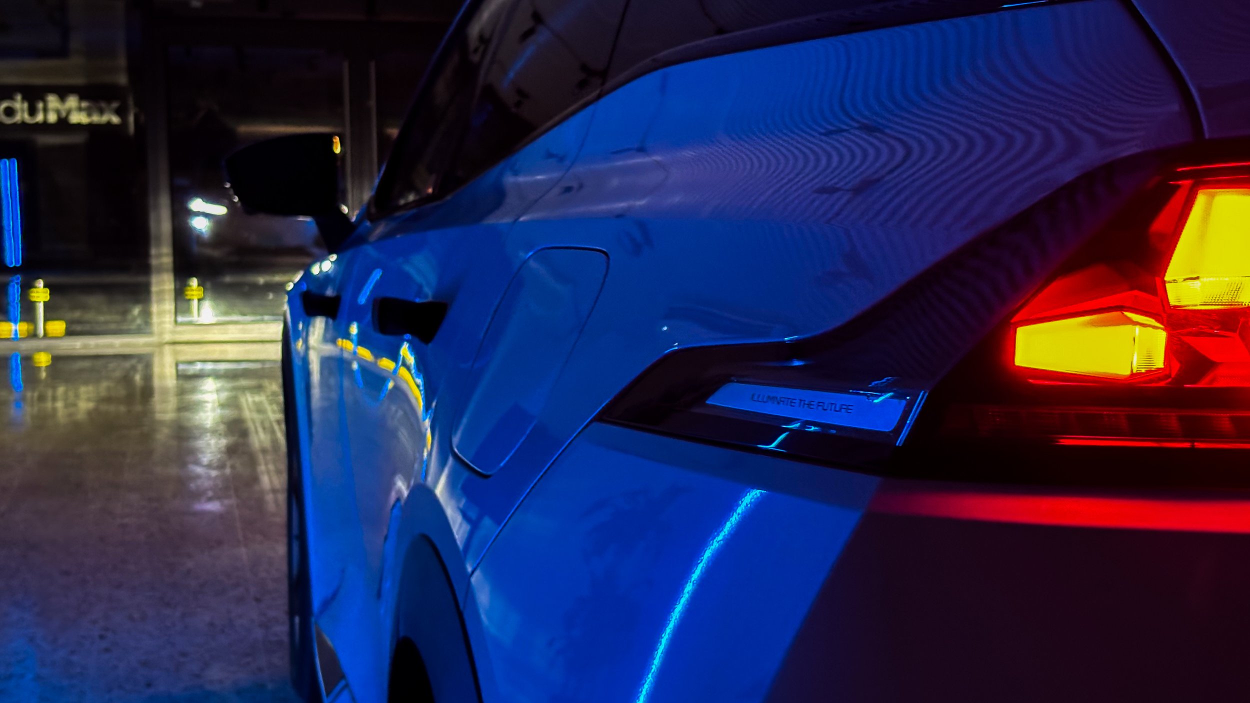 Close-up of a modern electric car with illuminated rear lights and a blue body, parked in a dimly lit urban area at night.