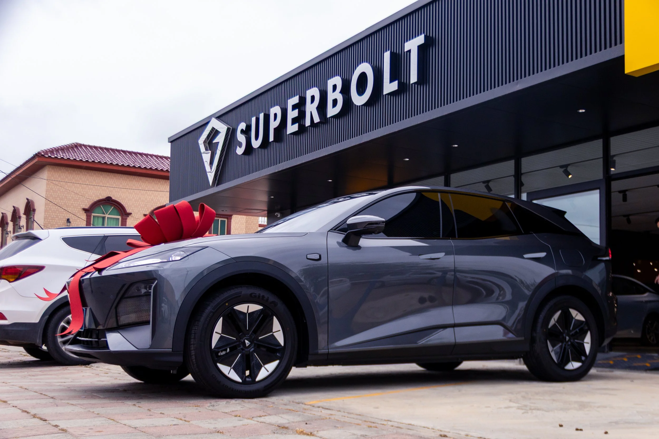 A new car with a red bow on the hood, parked in front of a dealership named SUPERBOLT.