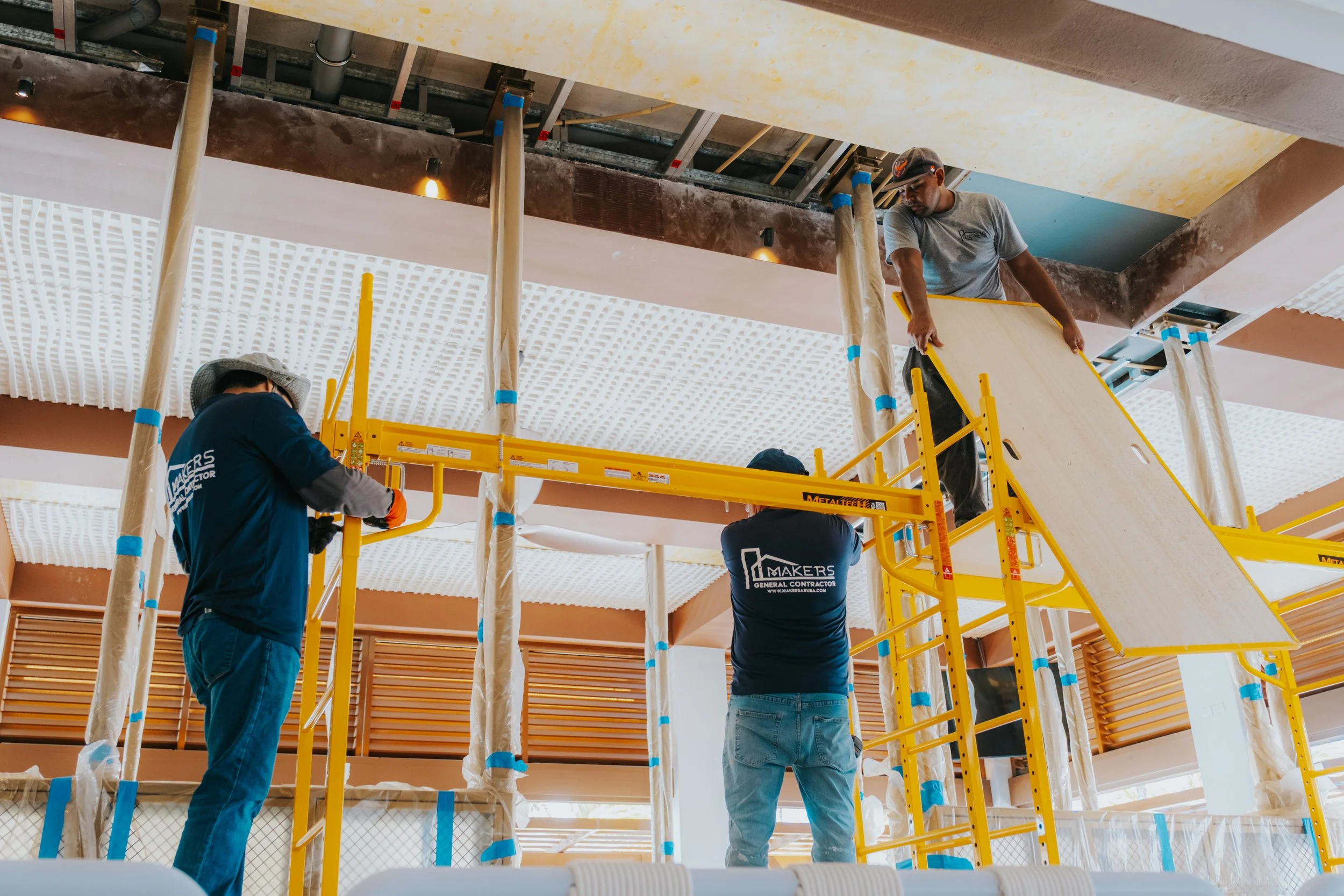 Workers installing ceiling panels on scaffolding in an indoor space, with some wearing uniforms and moving equipment.