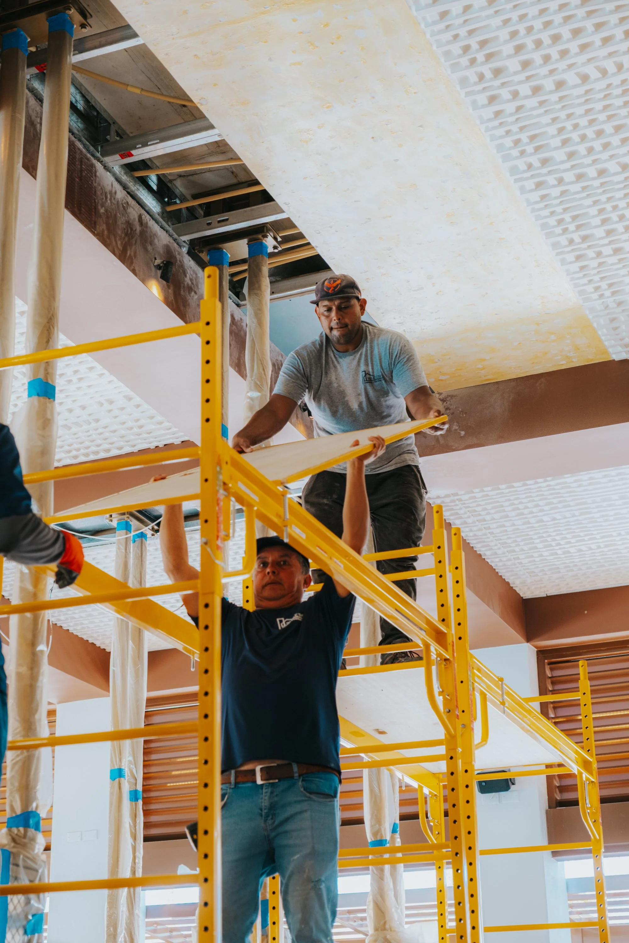 Construction workers installing ceiling panels, with scaffolding and construction materials visible.