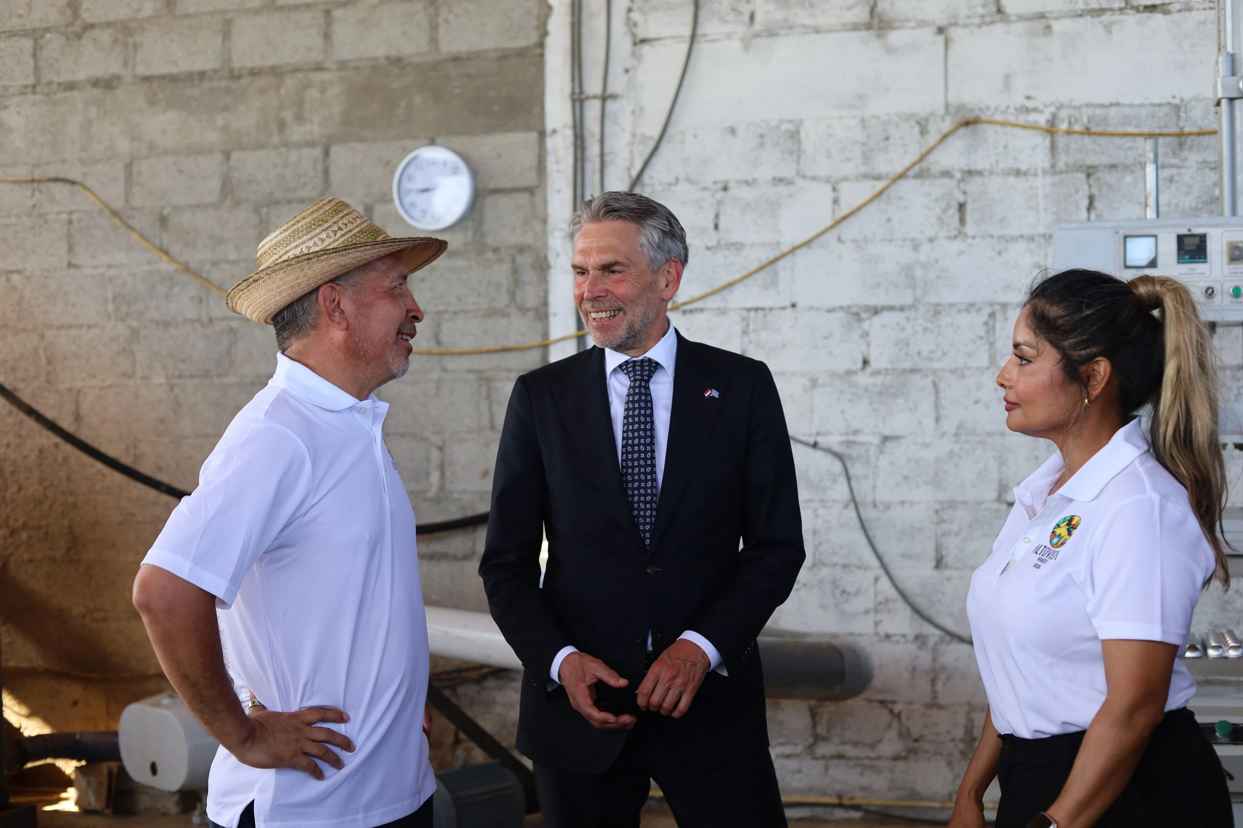 Three people engaged in conversation in an industrial setting with brick walls. The man on the left is wearing a white polo shirt and a straw hat, the man in the middle is dressed in a business suit, and the woman on the right is wearing a white polo