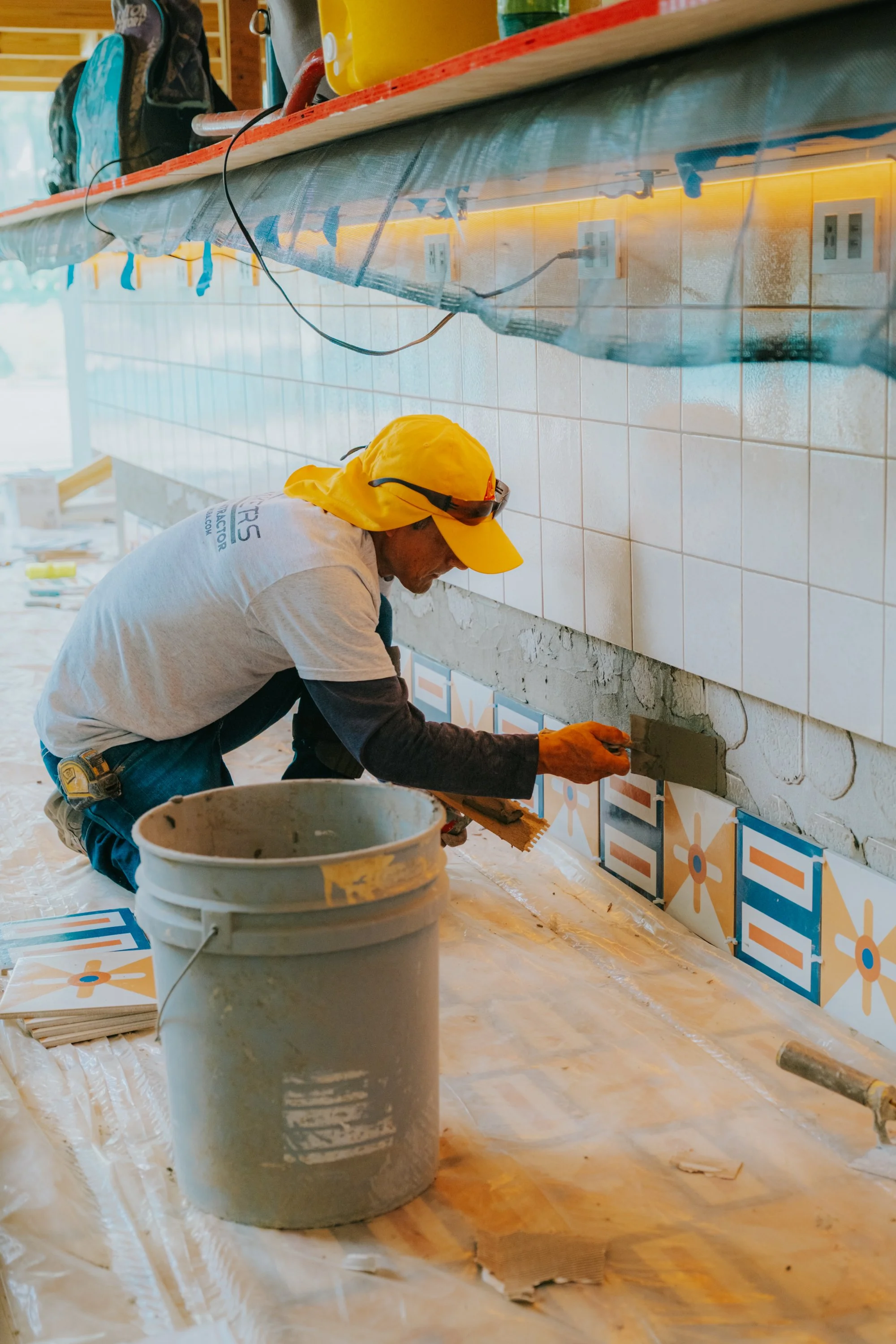 A construction worker wearing a yellow hat and gloves applies concrete to the base of a wall with decorative tiles, with construction tools and supplies nearby.