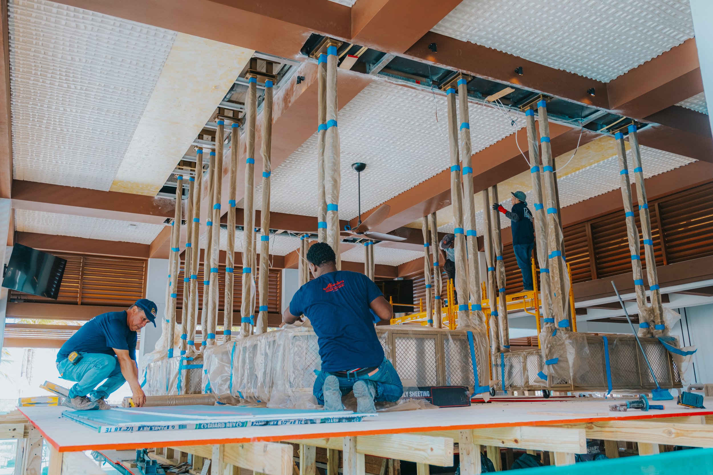 Construction workers installing ceiling panels inside a building. They are surrounded by scaffolding and construction materials.