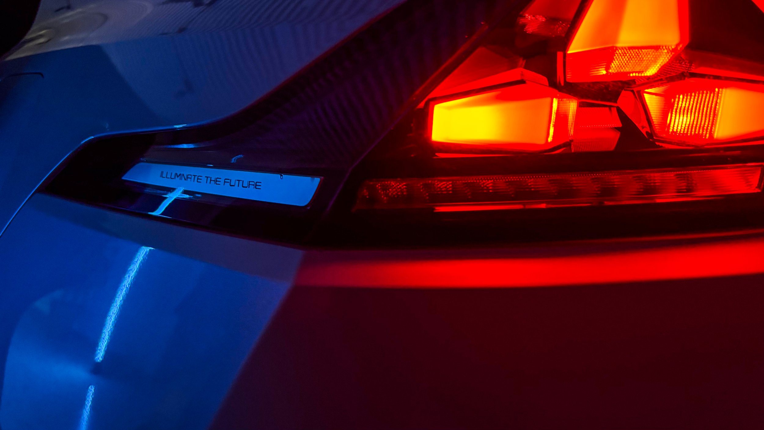 Close-up of the rear of a car with illuminated taillights and a blue accent light with the phrase 'ILLUMINATE THE FUTURE'.