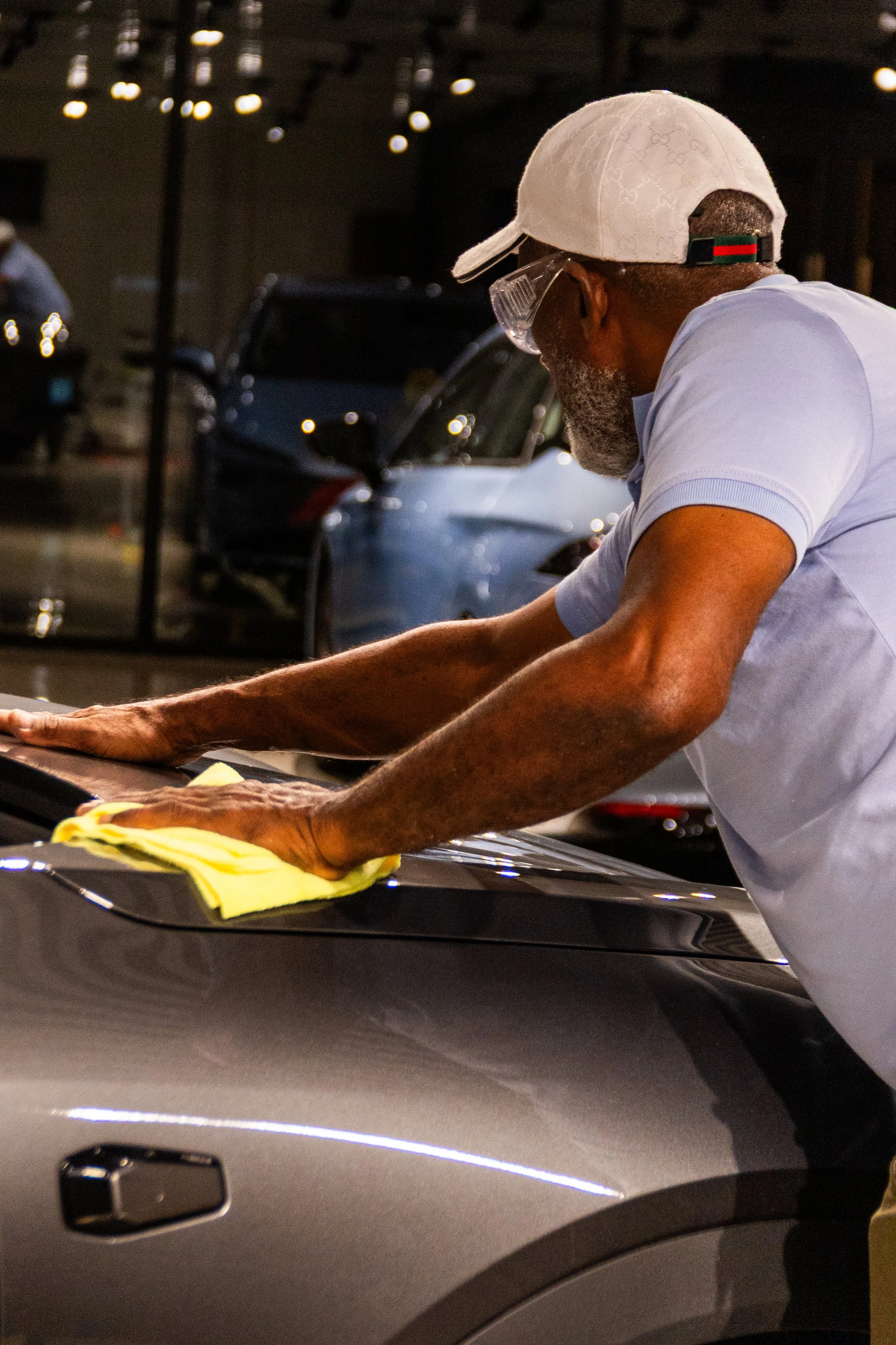 An elderly man polishing a car in a garage or showroom, wearing a white cap and glasses, with cars and reflections in the background.