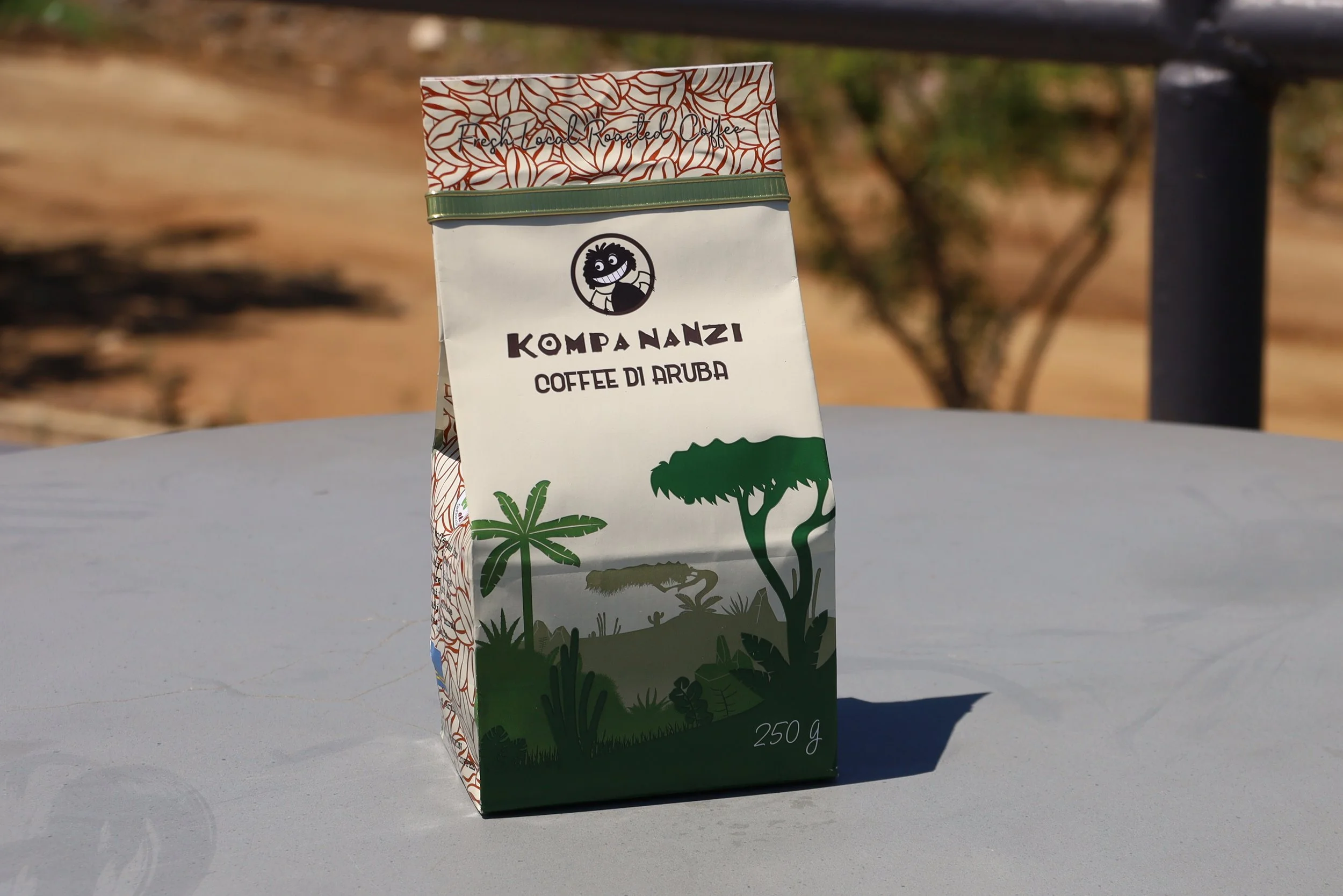 Coffee packaging with "Kompa Nanzi Coffee di Aruba" label, featuring green tropical tree illustrations on the front, on a gray table outdoors.