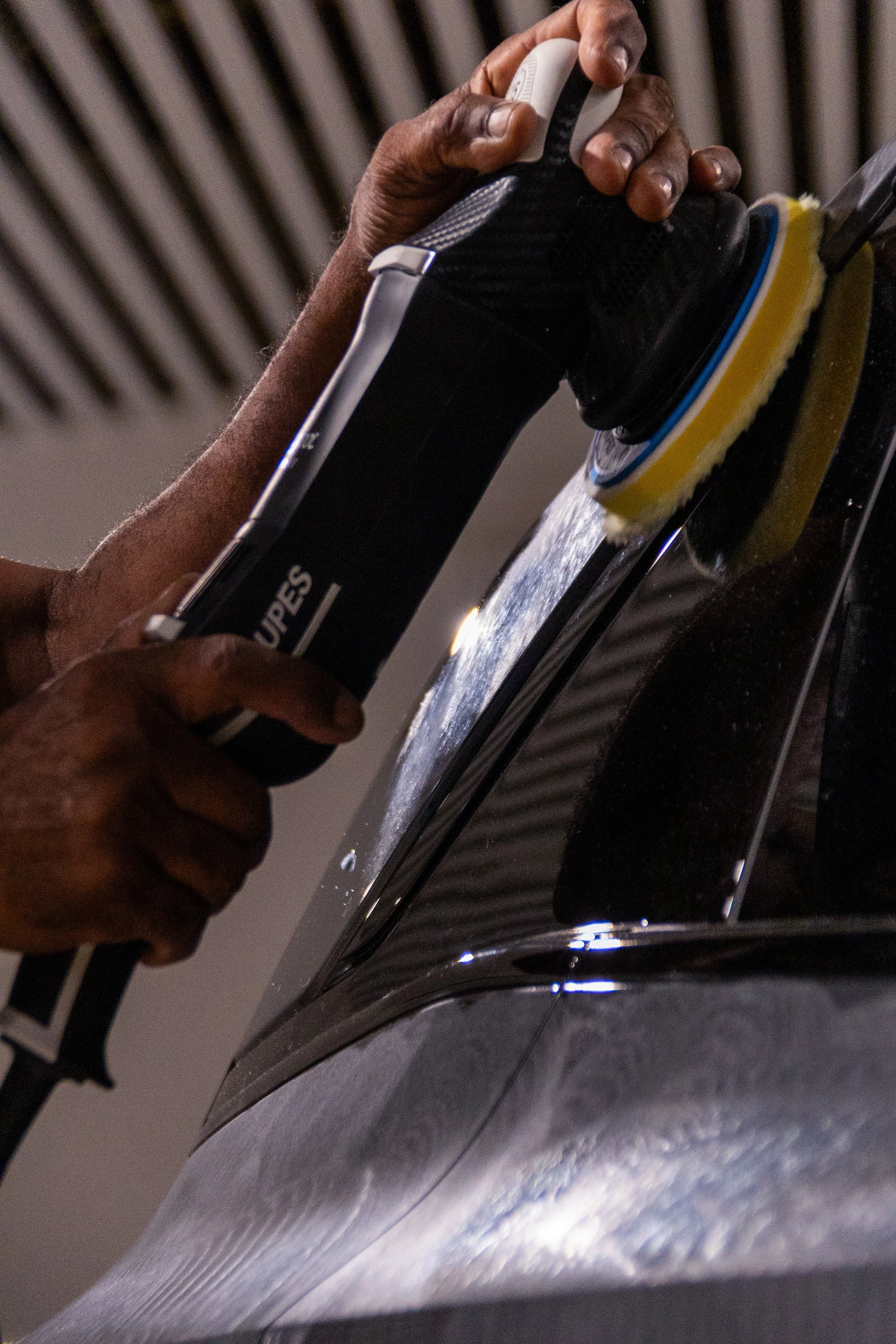 A person polishing a black car using a power buffer with a yellow pad.
